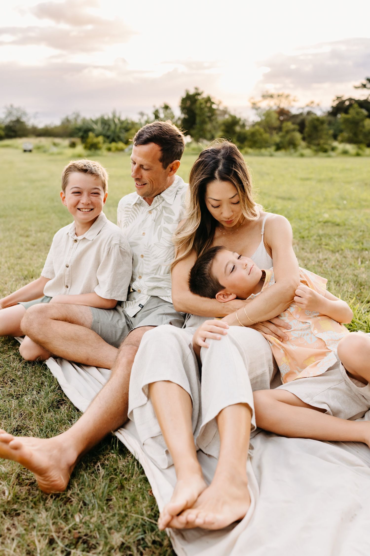 A group lounges together on a white blanket in a grassy field during a casual outdoor photoshoot.