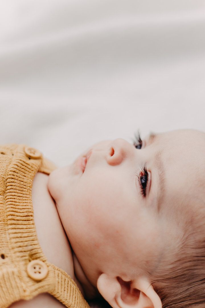 A baby wearing a yellow knit sweater lies on white bedding in a close-up profile view of eyelashes.