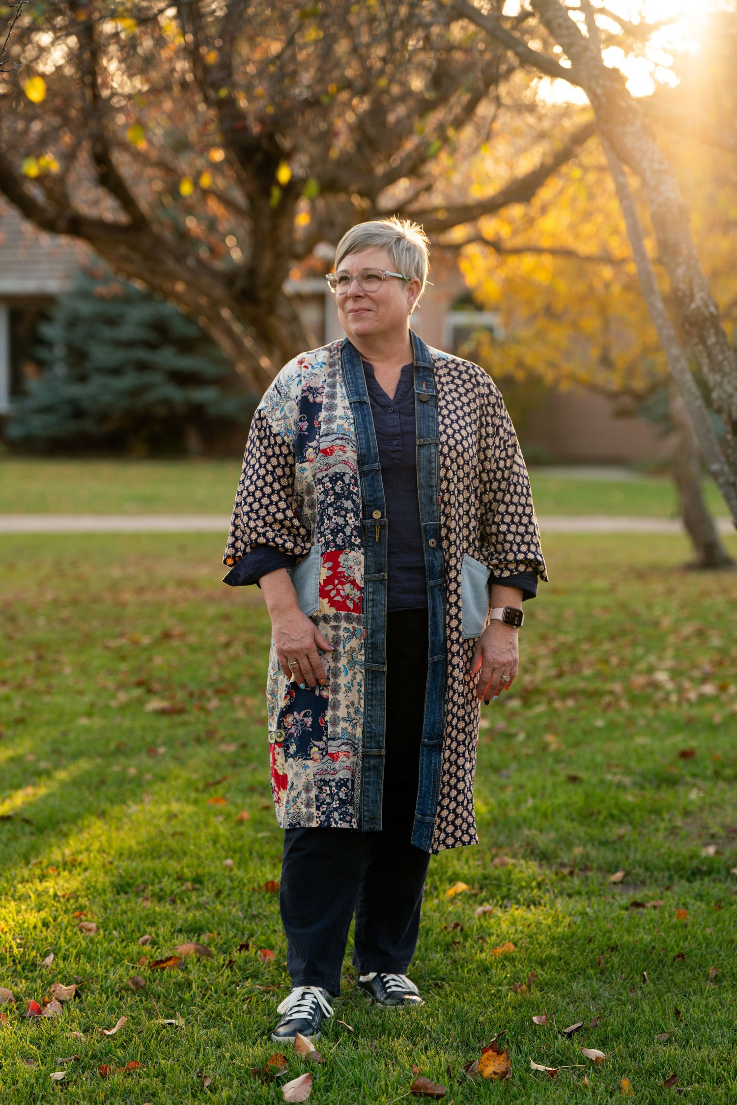 Person wearing a patterned jacket poses in a park with fall foliage and golden sunlight.