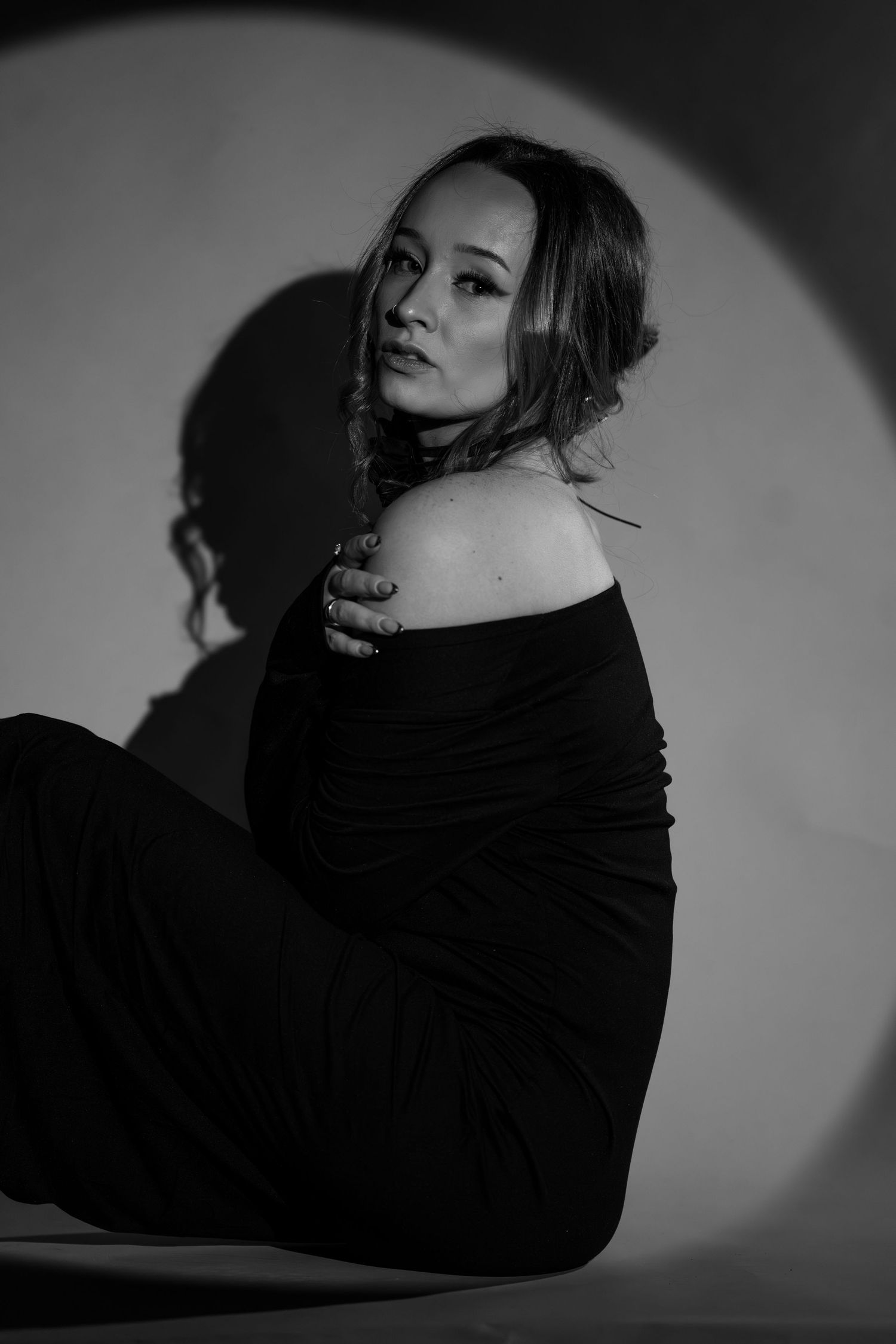 Artistic black and white photo of a woman in off-shoulder dress posing against a circular light backdrop.