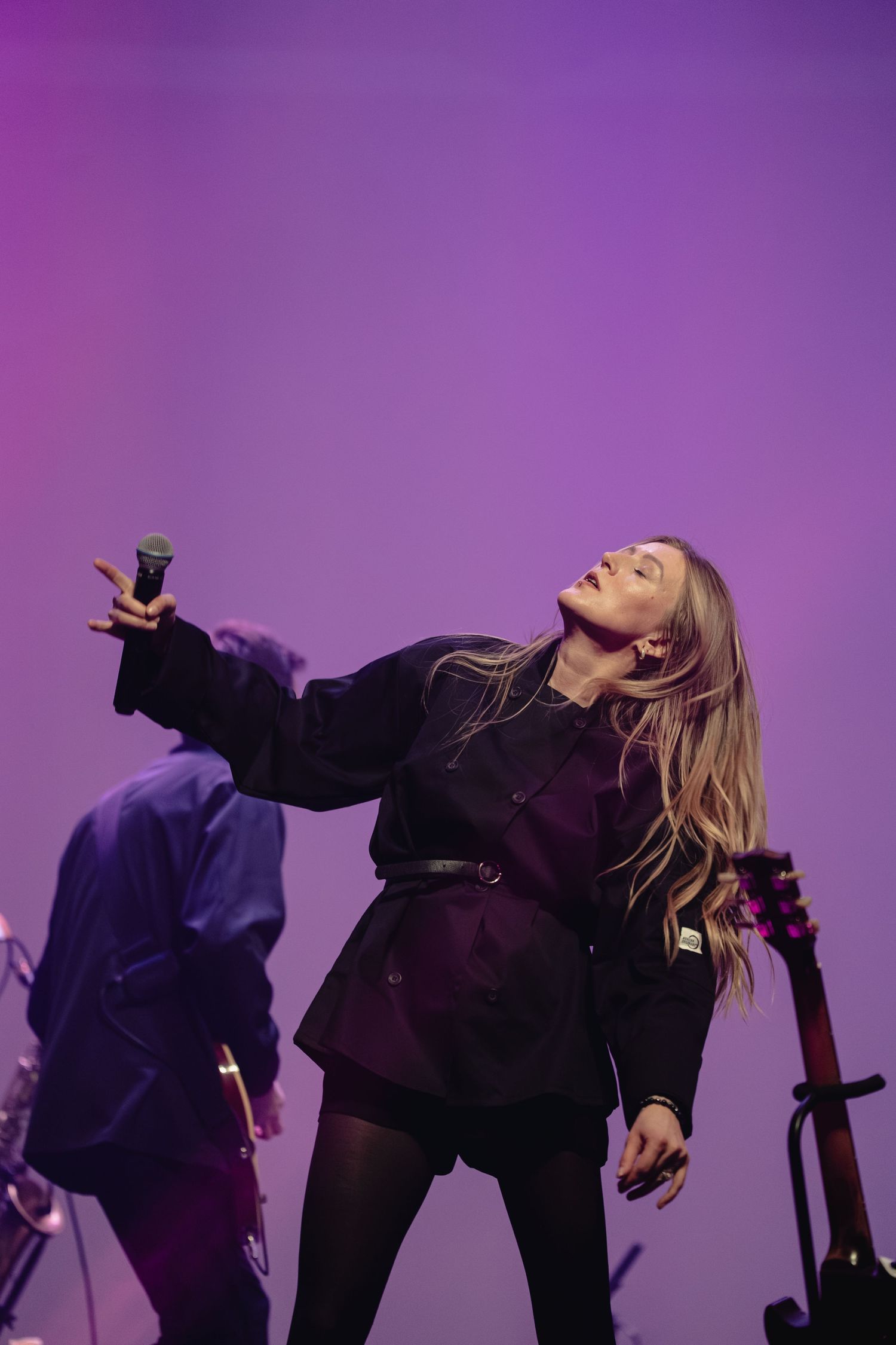 Concert performer in black outfit strikes dynamic pose against purple stage lighting.