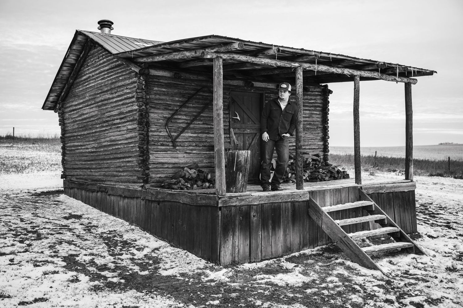 Series of weathered wooden cabins in a snowy rural landscape captured in black and white.