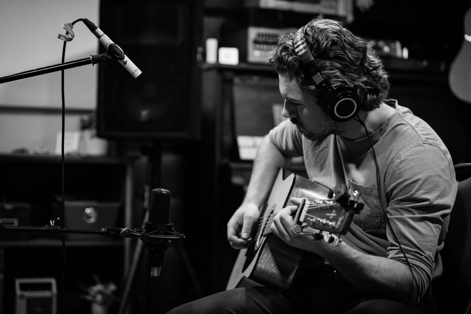 Black and white photo of musician recording with acoustic guitar and headphones in studio setting.