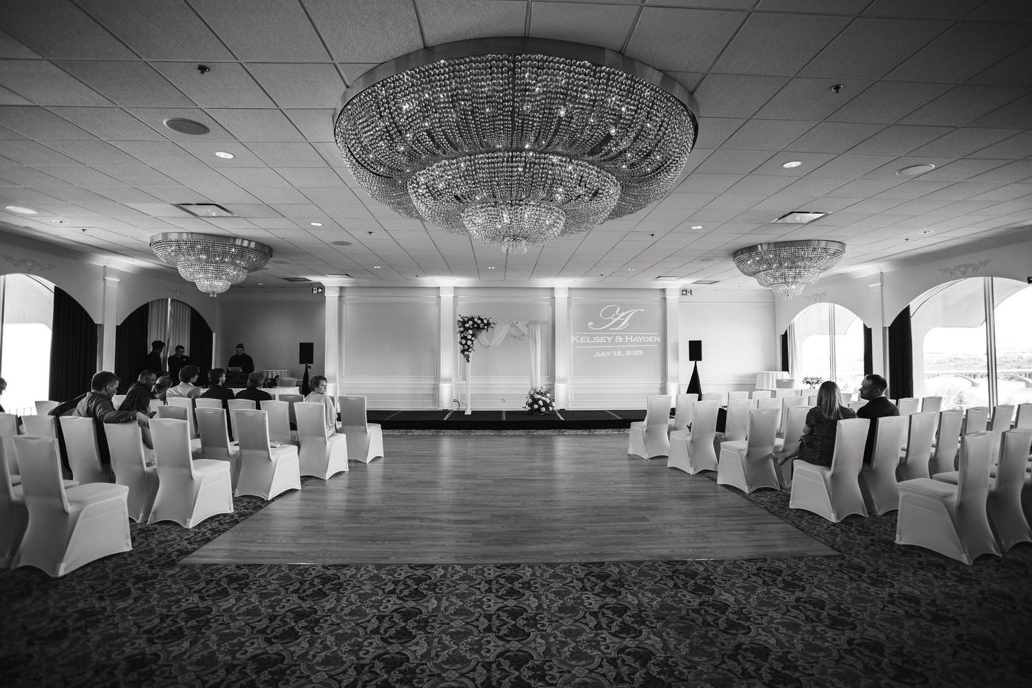 Elegant ballroom with crystal chandeliers and white chair covers set for wedding ceremony.