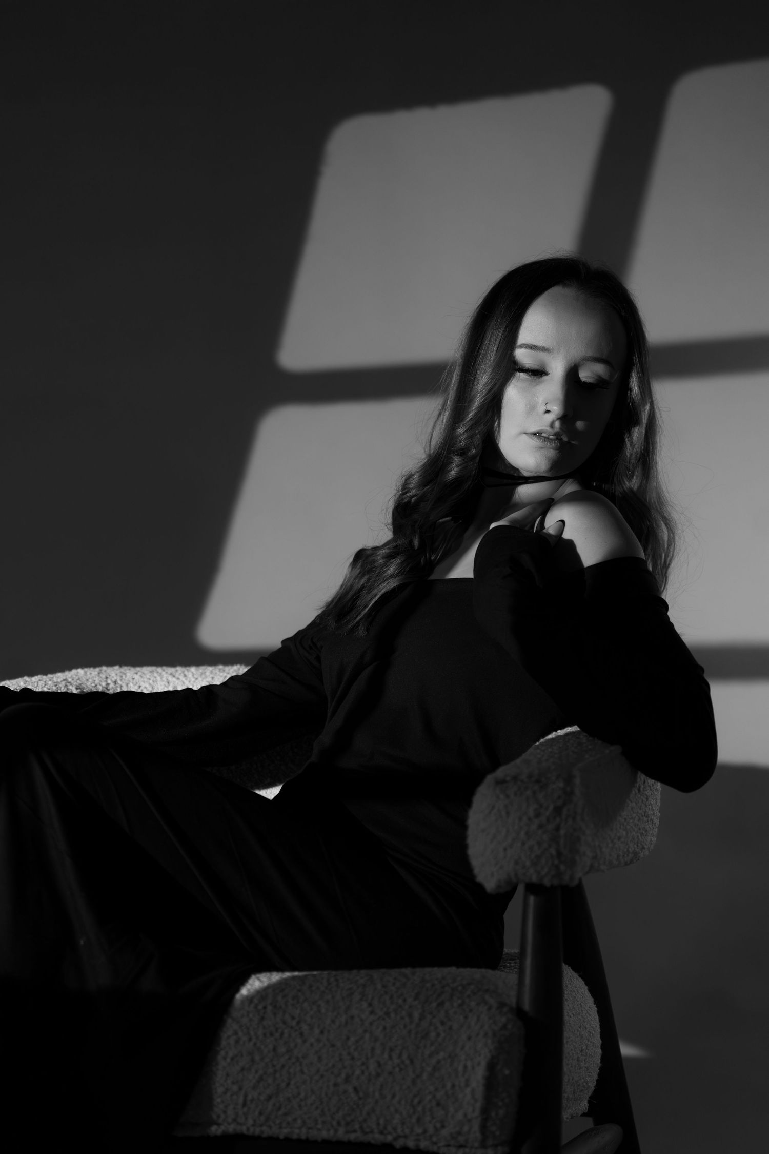Atmospheric black and white photo of a woman in dark clothing sitting in chair with window shadows.
