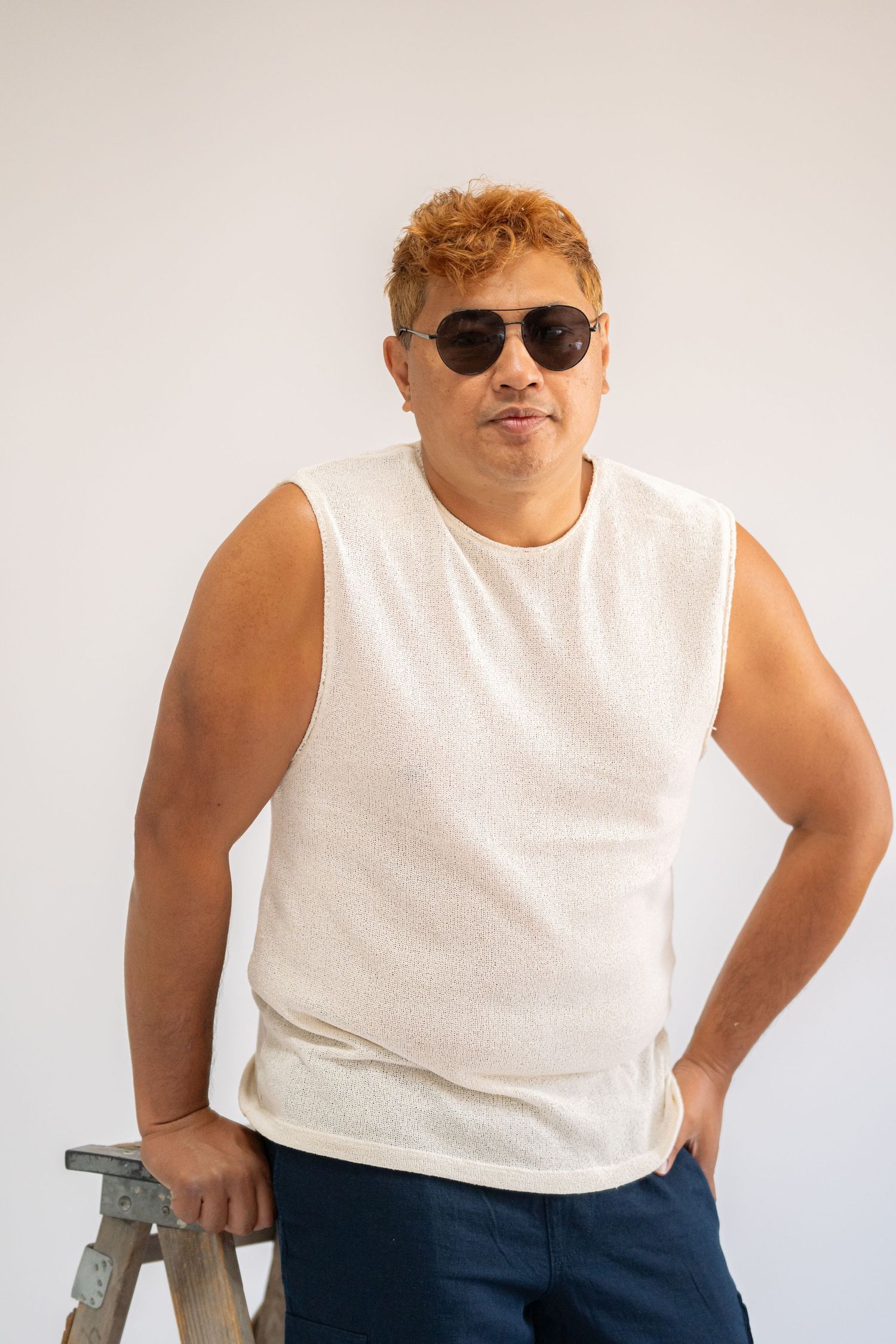 Person in white sleeveless shirt and dark pants posing against light background.