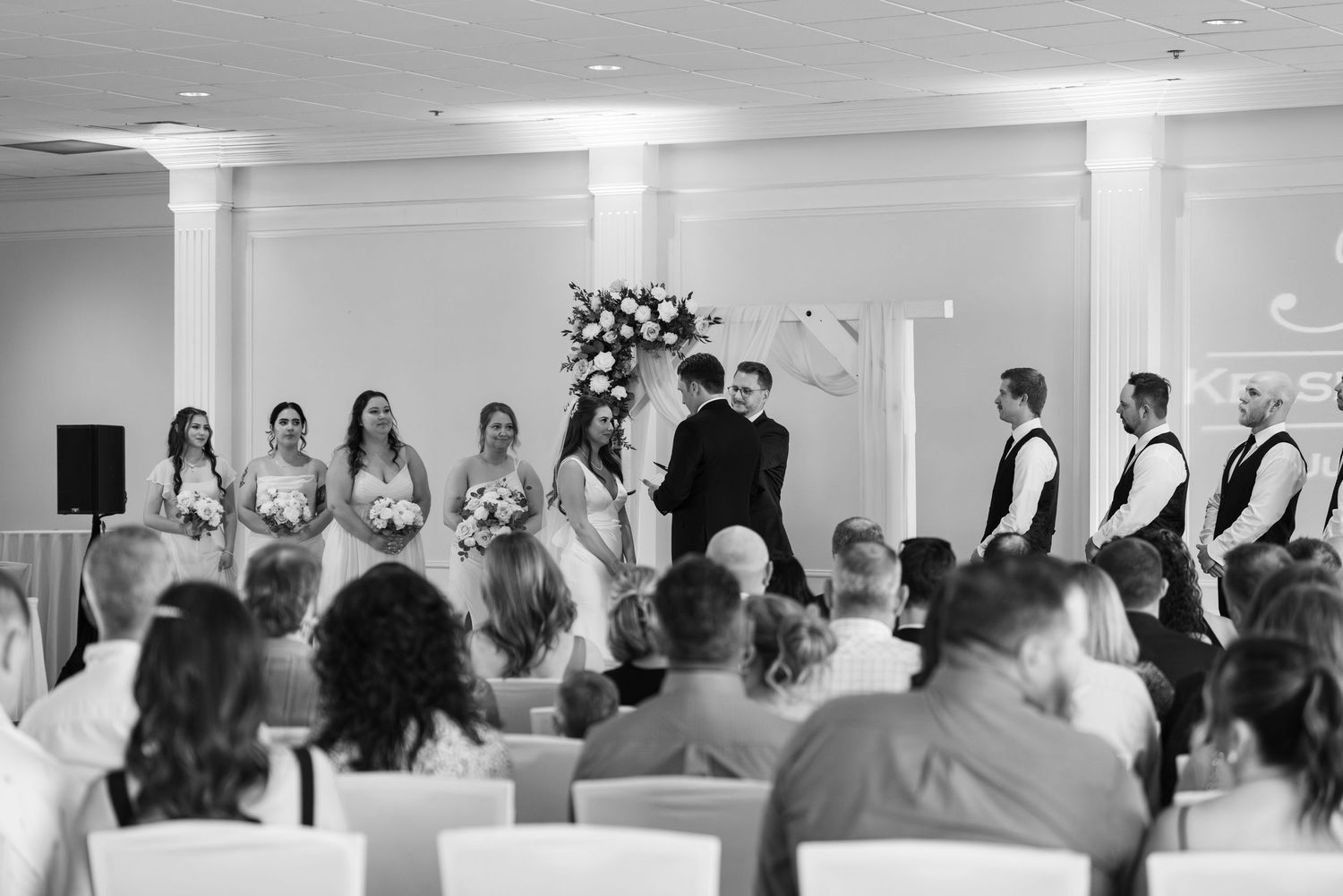 Wedding ceremony in progress under elegant chandeliers with floral arch.