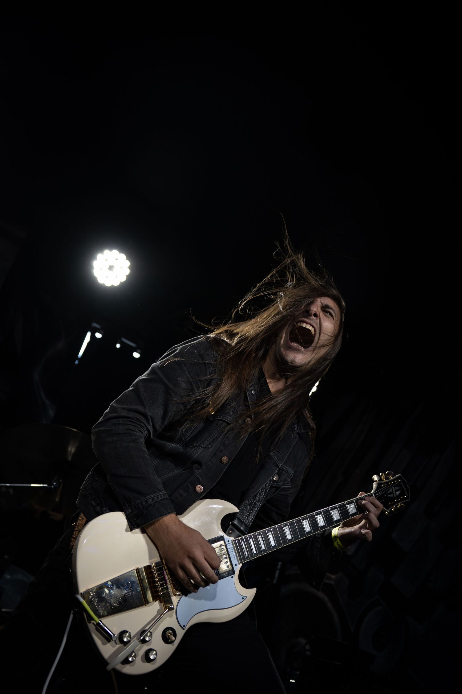 Guitarist performs energetically on stage with a white electric guitar against dark background.