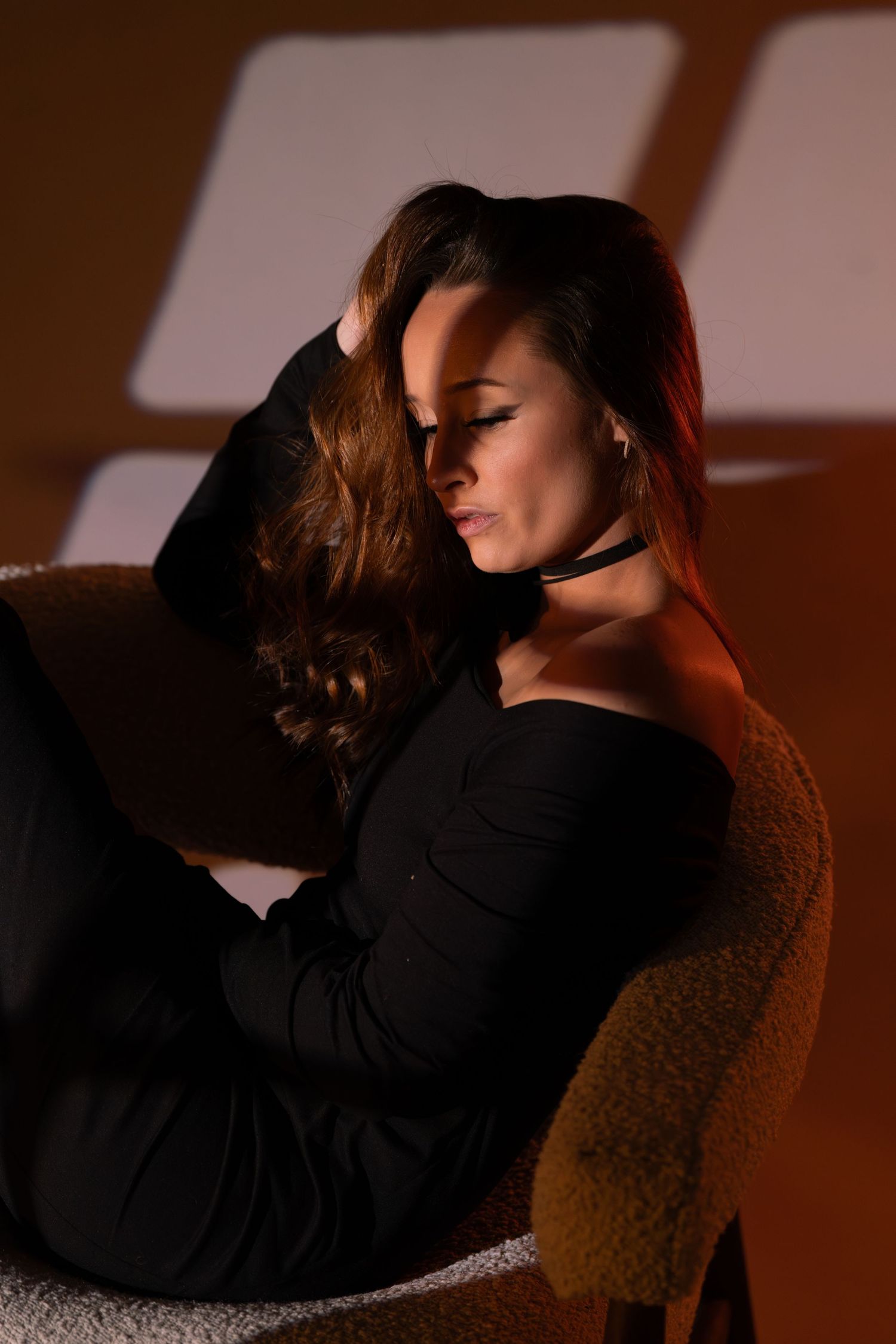Moody portrait of a woman in black dress reclining on a chair with warm lighting and shadows.