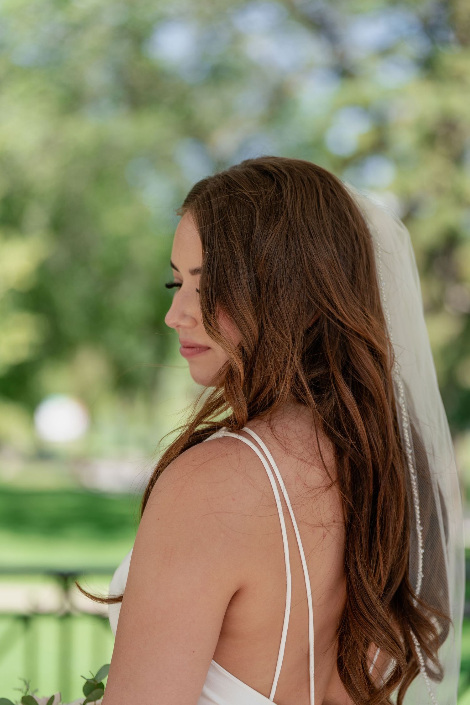 Side profile of bride in sleeveless wedding dress with veil against lush green garden backdrop.