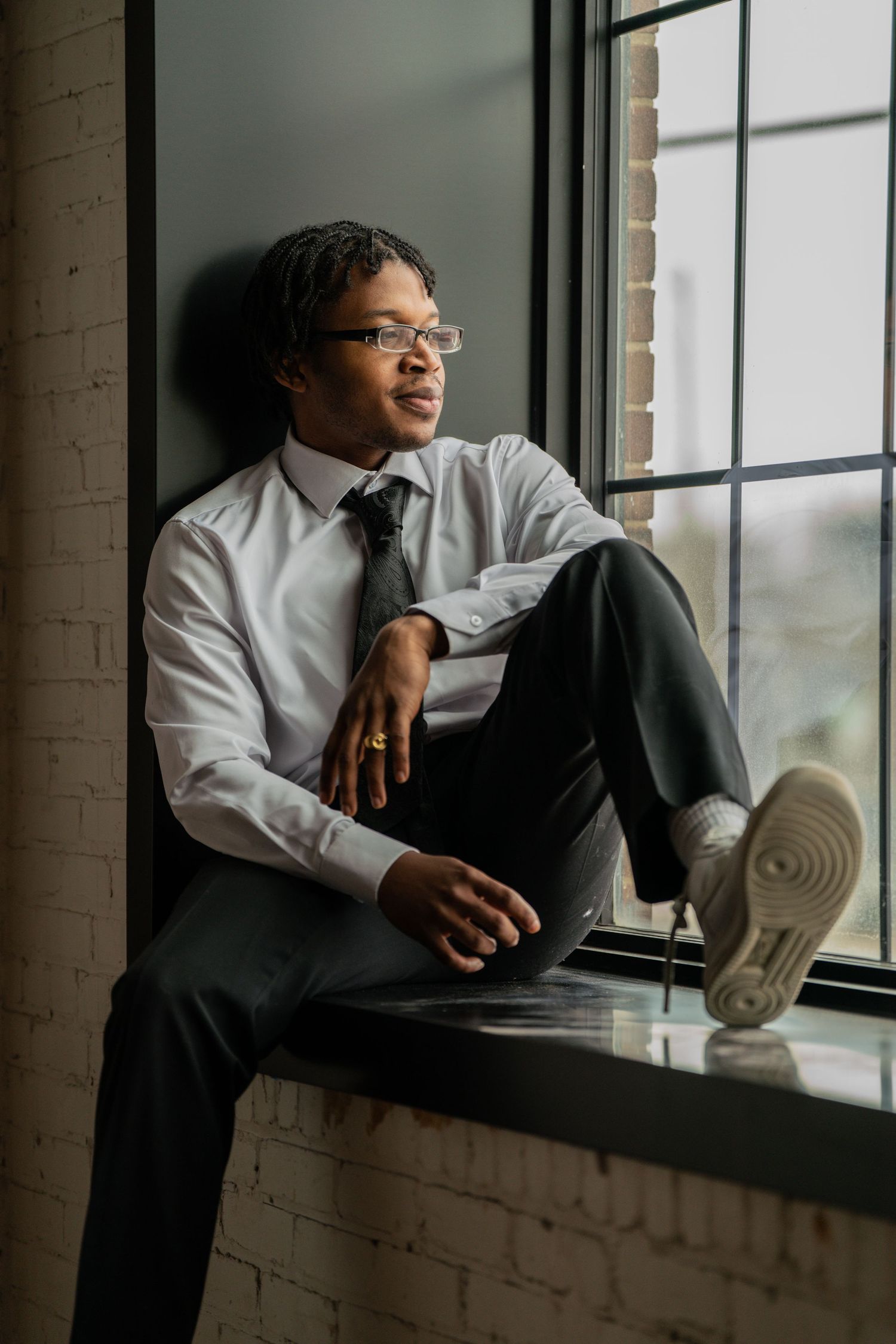 Someone in formal wear gazes thoughtfully through a bright window in a brick room.