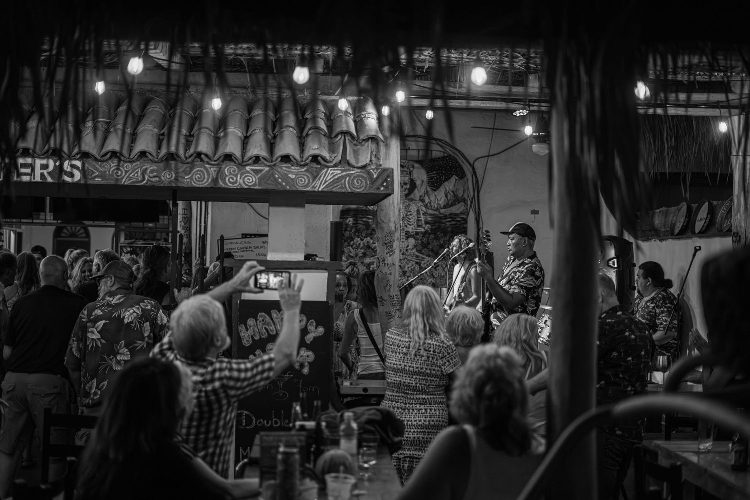 Black and white image of crowded bar scene with people gathered under string lights.