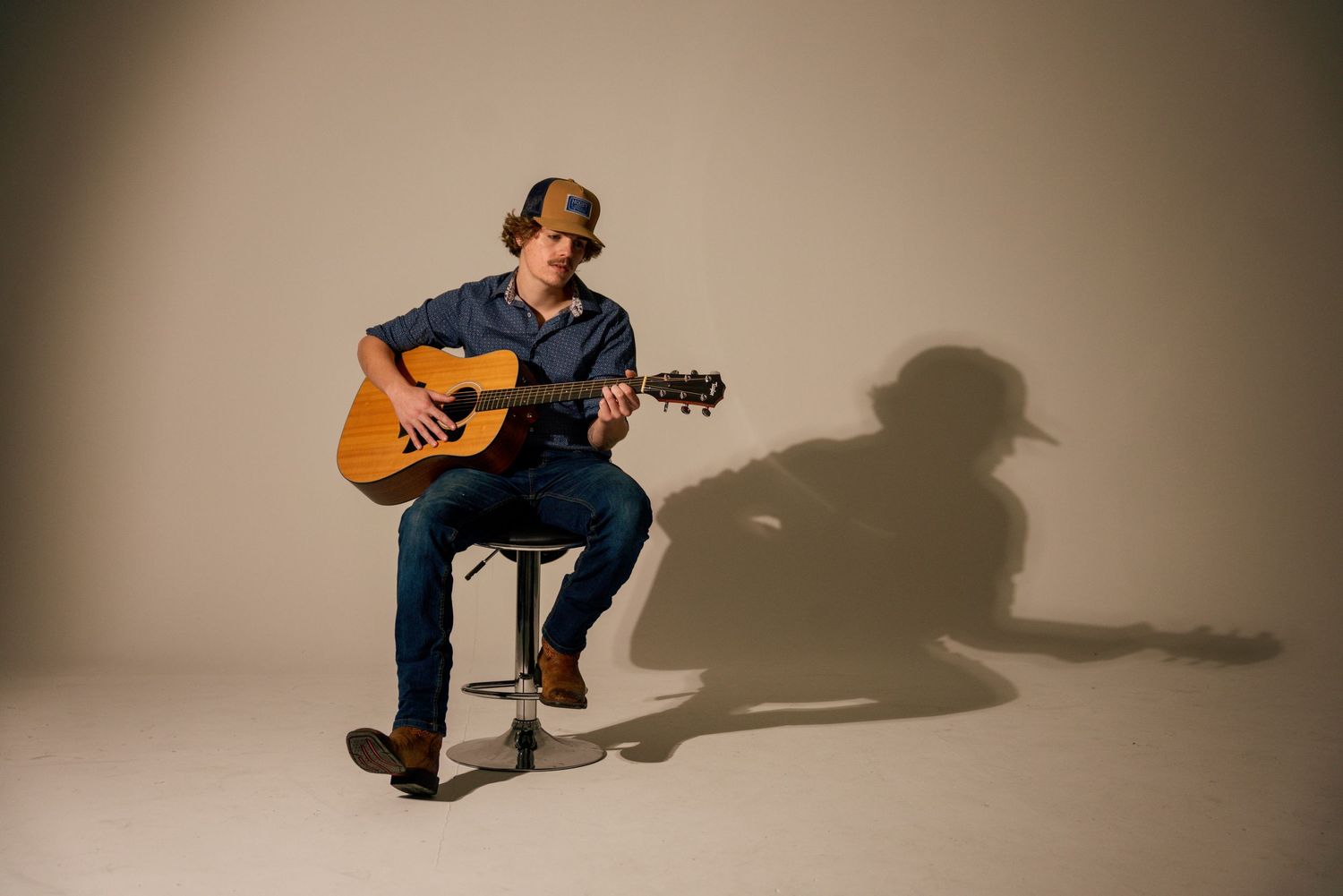Musician playing acoustic guitar while sitting on a stool casting dramatic shadow against light background.