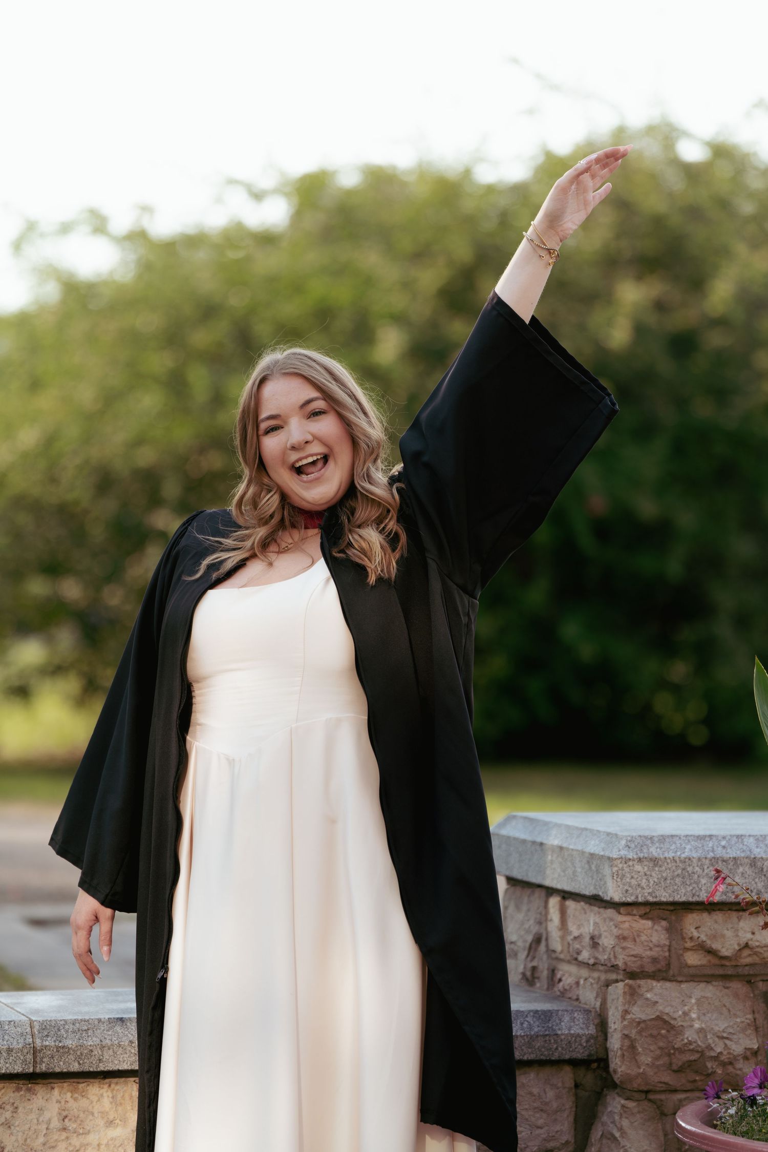 A graduate raises arm in celebration wearing graduation gown over white dress.