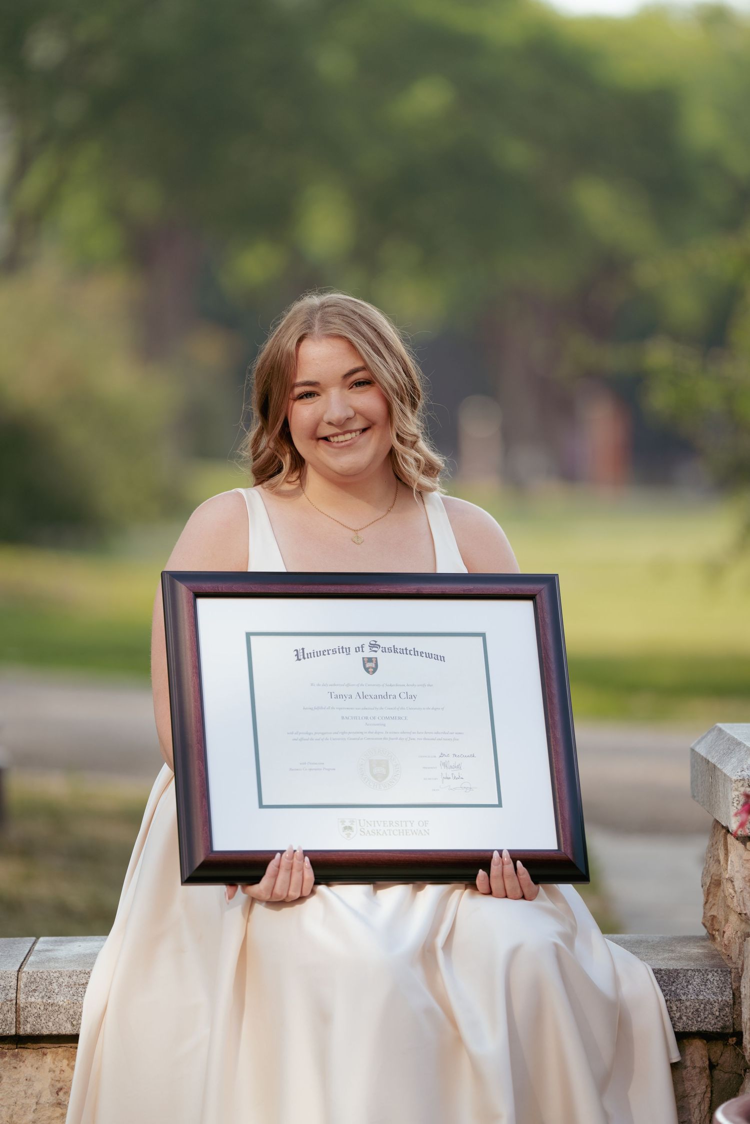 A graduate displays university diploma while sitting on stone wall in white dress.