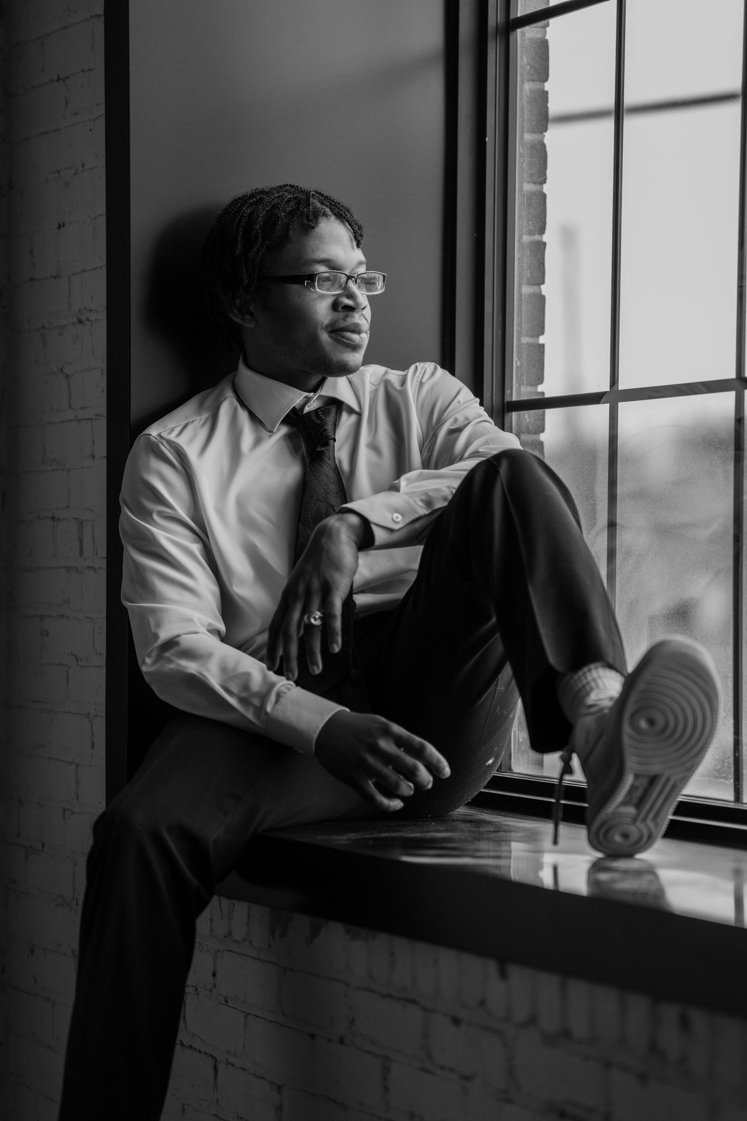 A person in business attire sits contemplatively by a large window in black and white.