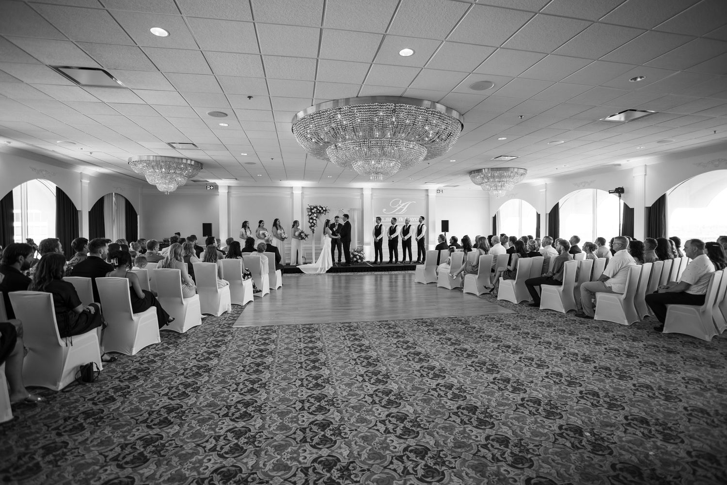 Black and white photo of wedding venue with rows of chairs and grand chandeliers.