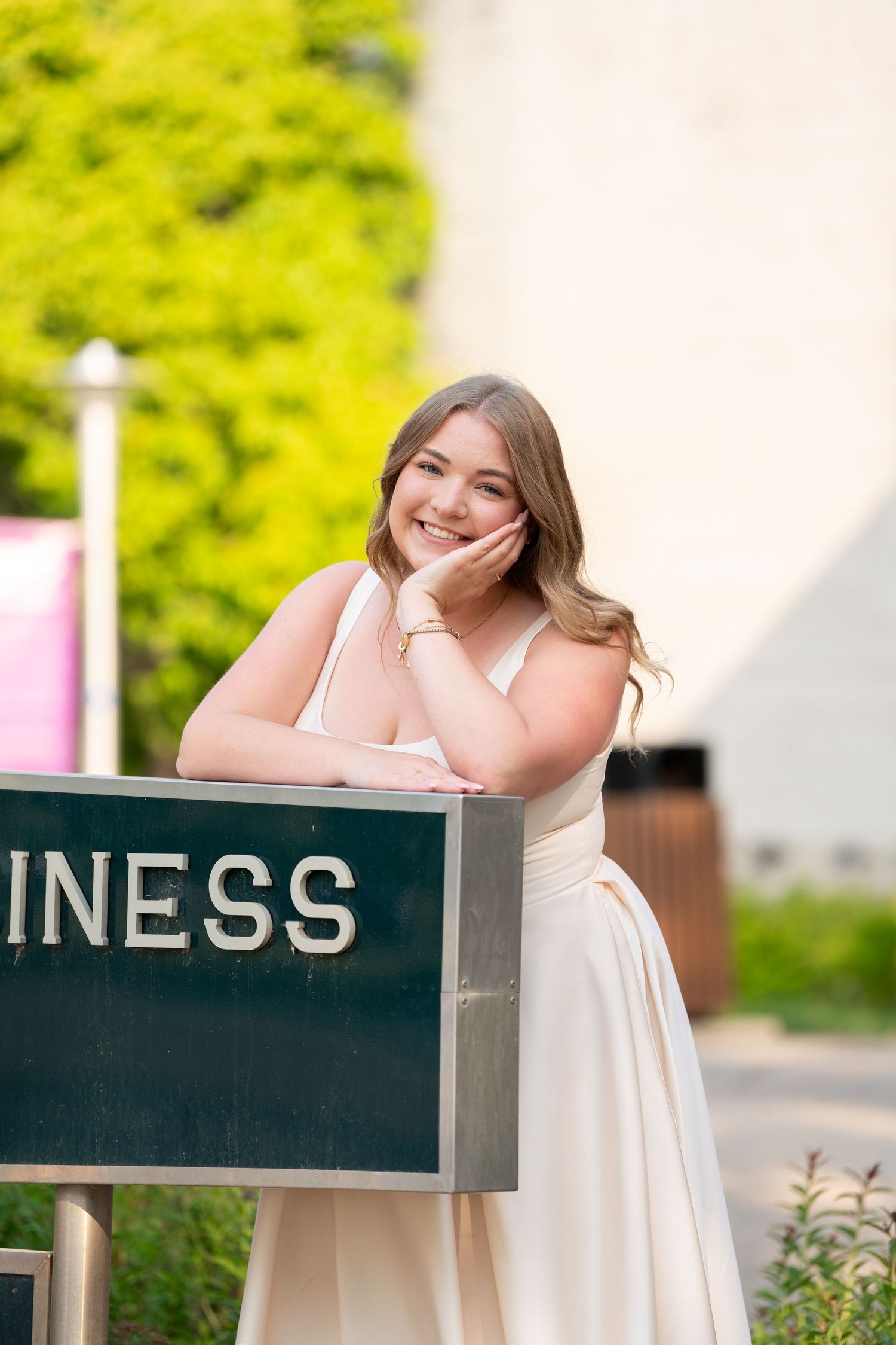 A graduate in white dress leans on a business sign during graduation photoshoot.