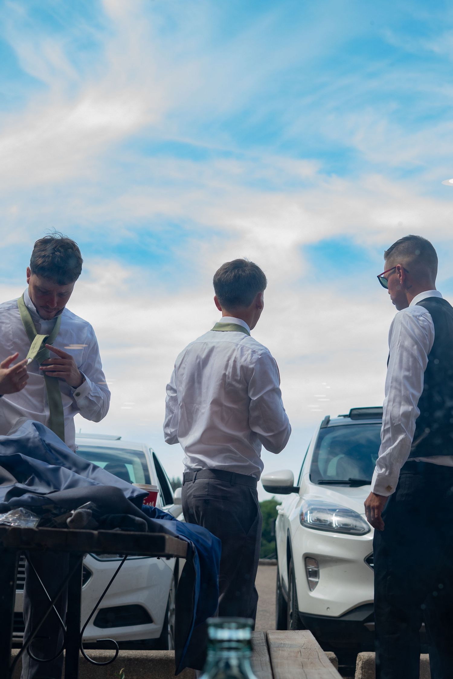 Group in formal attire gathered outdoors under blue sky near parked cars.
