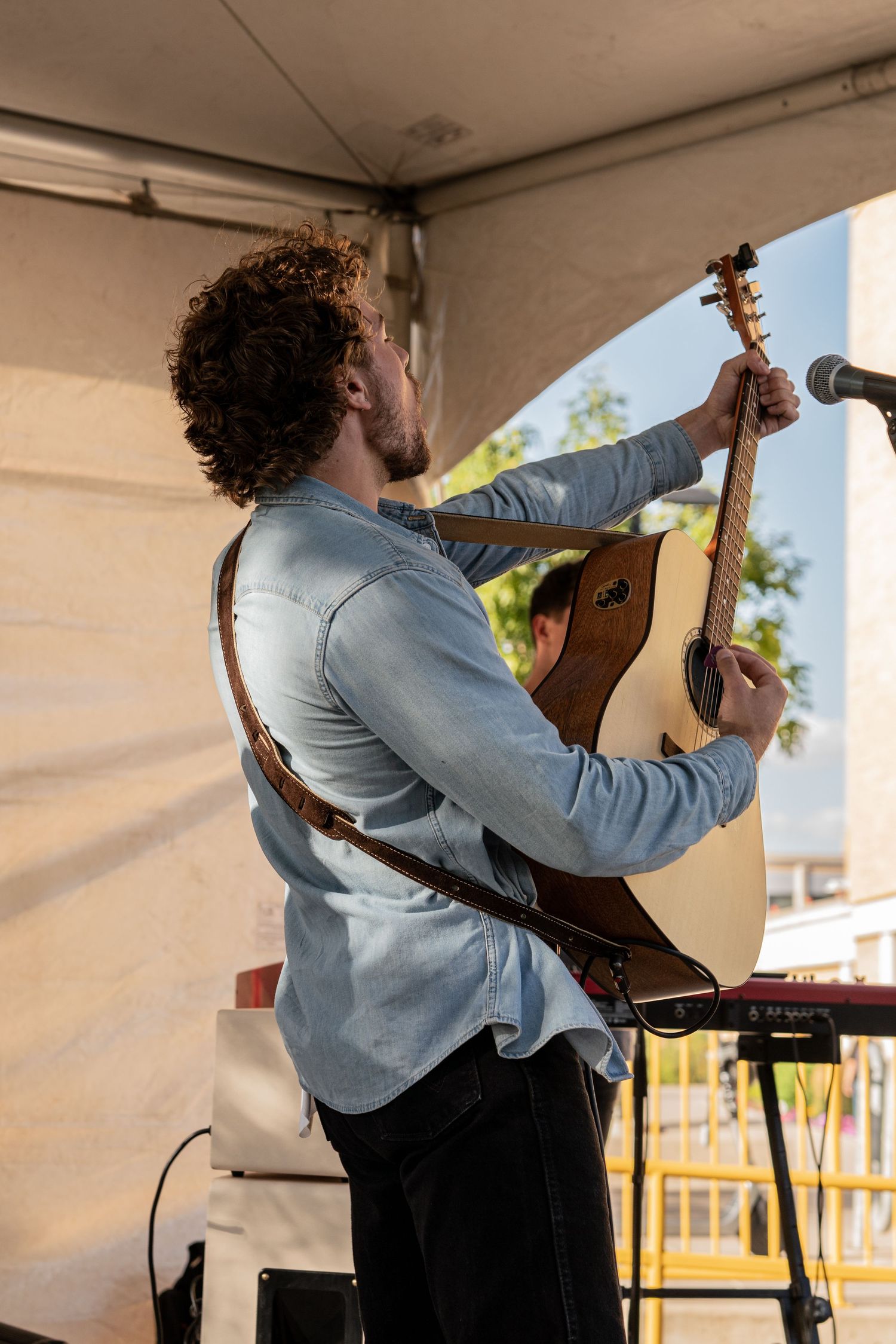 Musician playing acoustic guitar under outdoor tent during daytime performance.