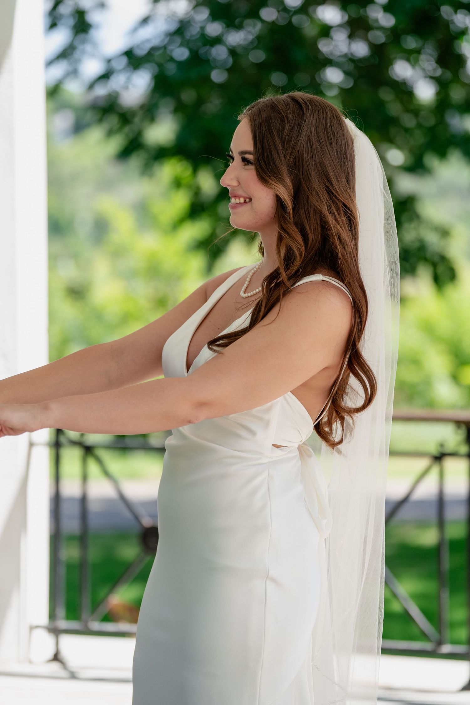 Bride in a white wedding dress with flowing veil poses gracefully on a sunny porch with green foliage in background.