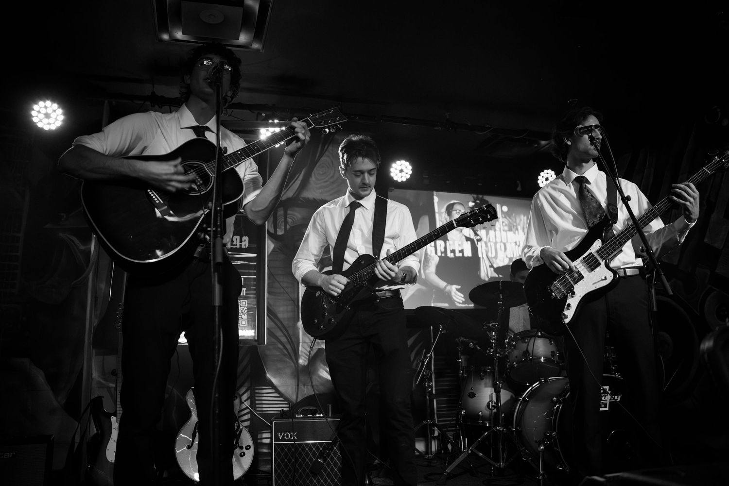 Black and white concert photo showing band performing with guitars and drums on stage.