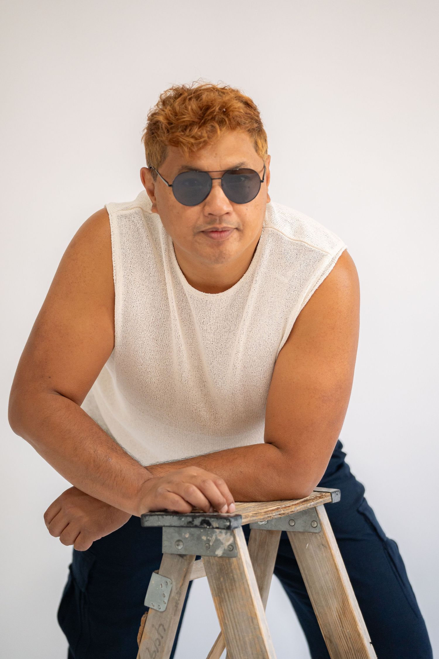 Portrait of a person in sleeveless white shirt sitting on wooden stool wearing aviator sunglasses.