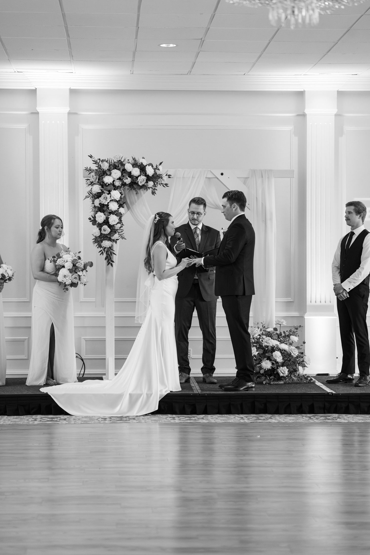Wedding ceremony taking place under floral arch in elegant white ballroom.