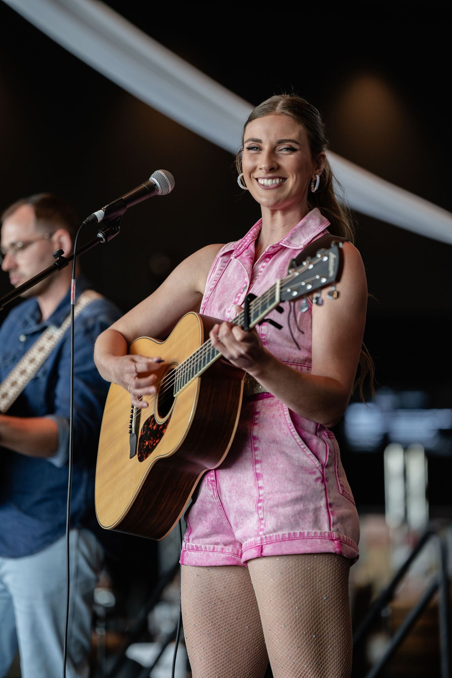 Musicians performing on outdoor stage with acoustic guitars and microphones.