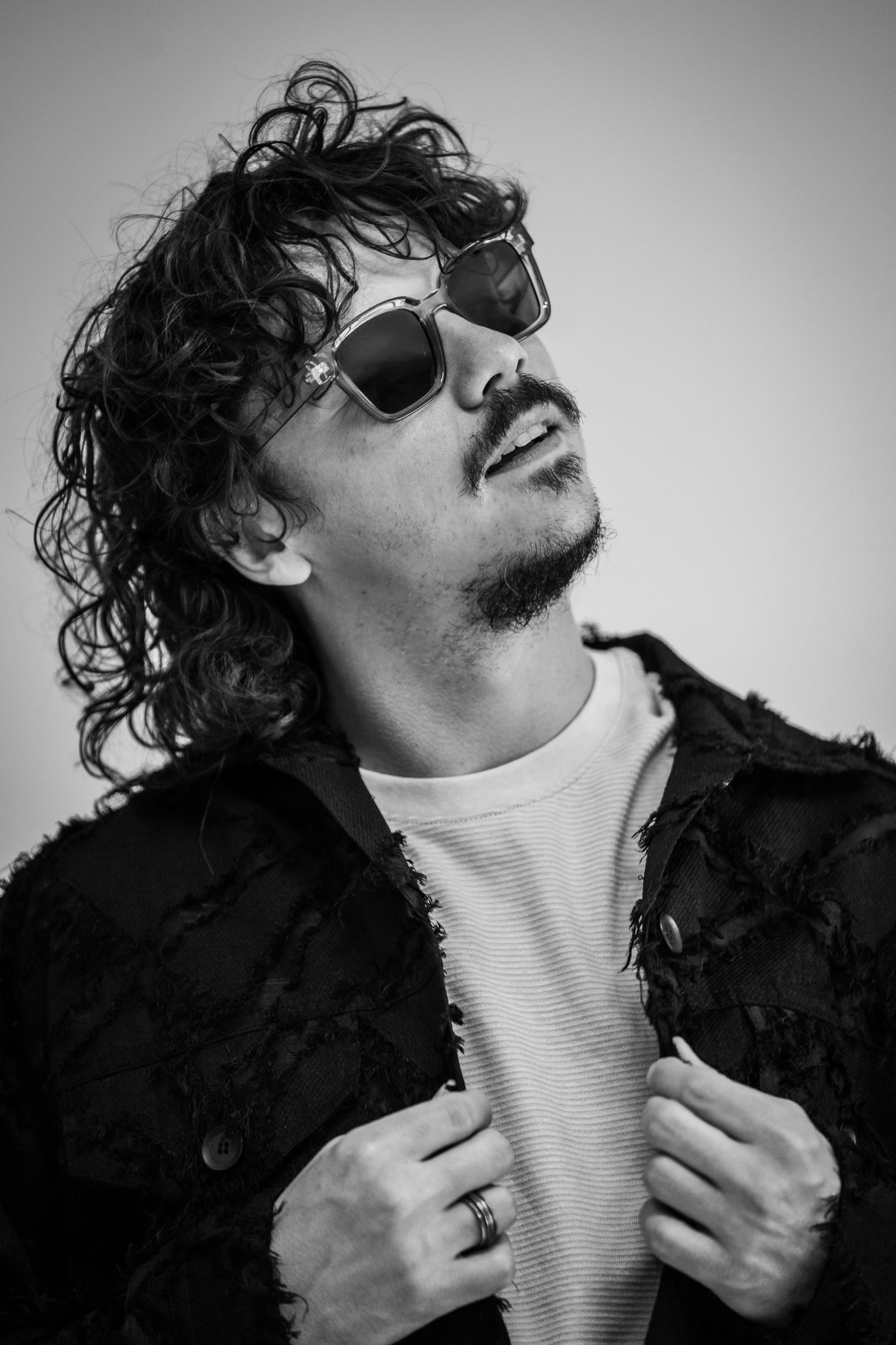 Dramatic black and white photo of a person in sunglasses and textured jacket with curly hair.
