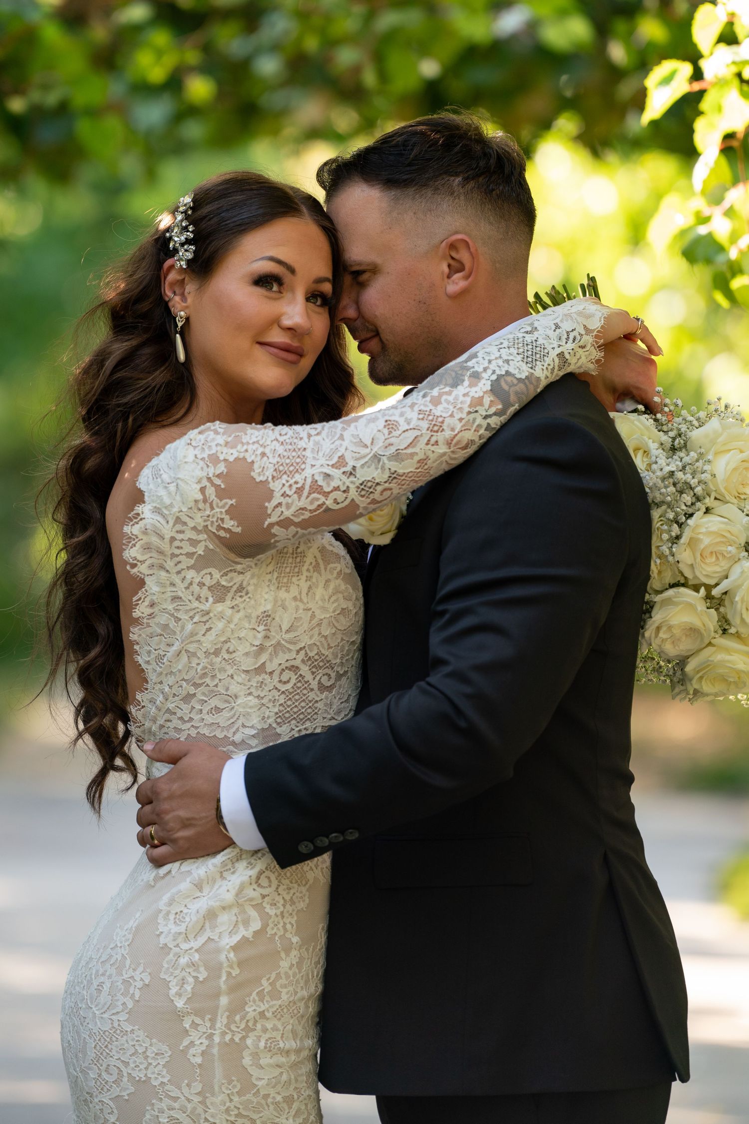 Romantic wedding portrait of couple in lace gown and black suit embracing in garden setting.