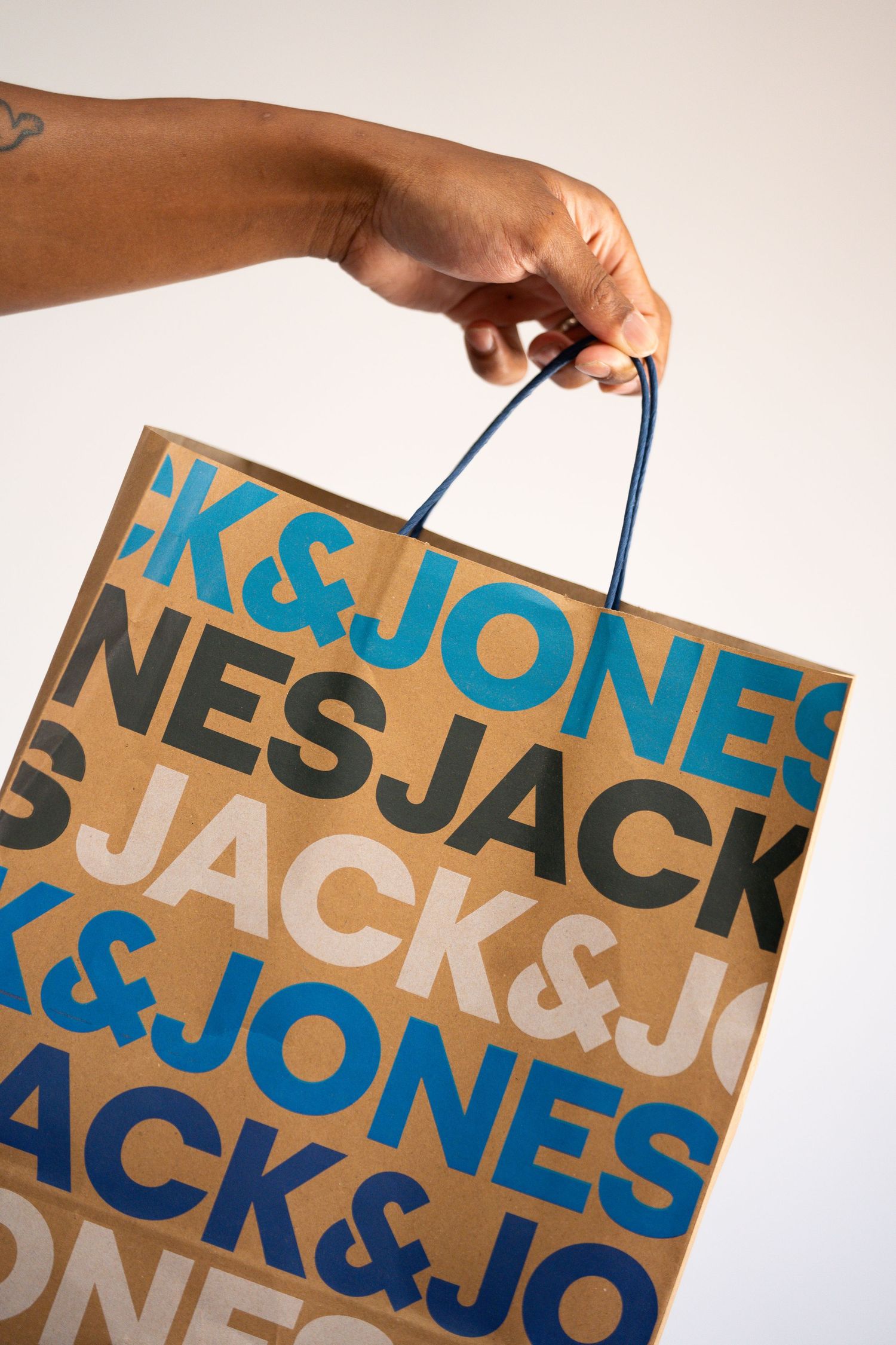 Hand holding brown paper shopping bag with repeating Jack & Jones text pattern.