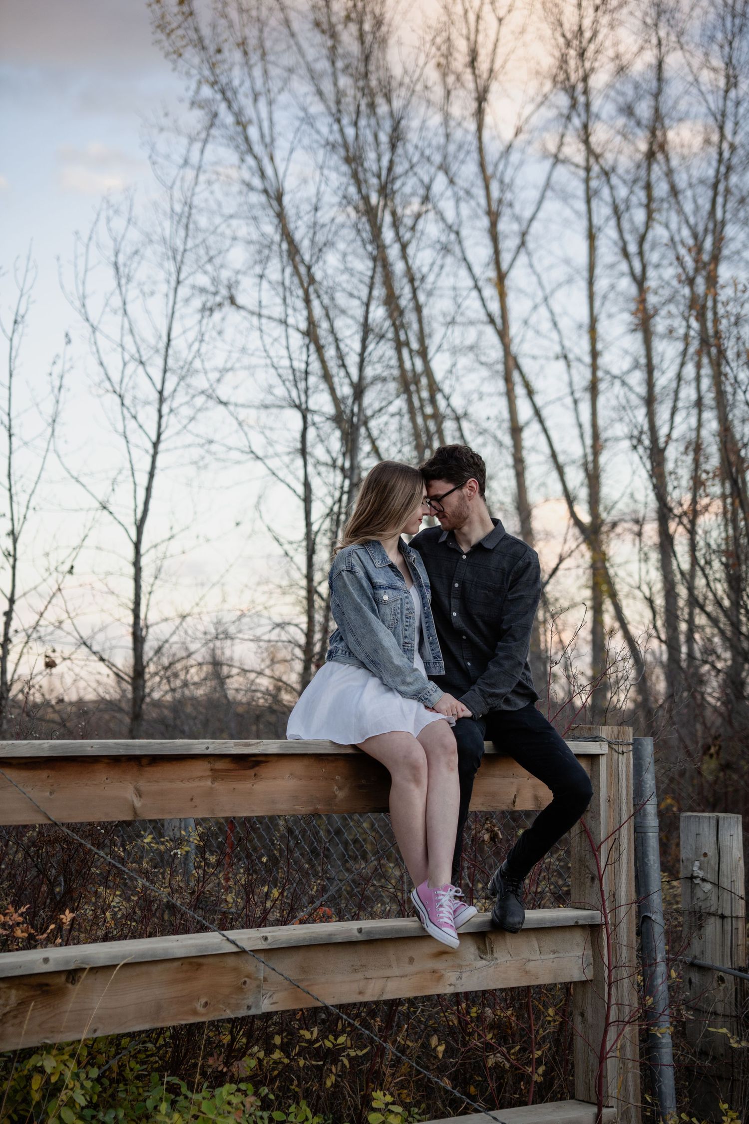 Couple sitting close together on wooden fence during sunset with bare trees in background.