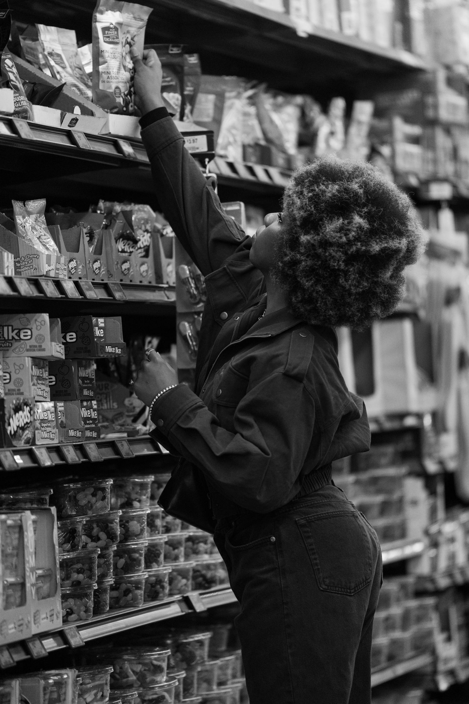 Person in black jacket reaching for items on high grocery store shelf in black and white photo.