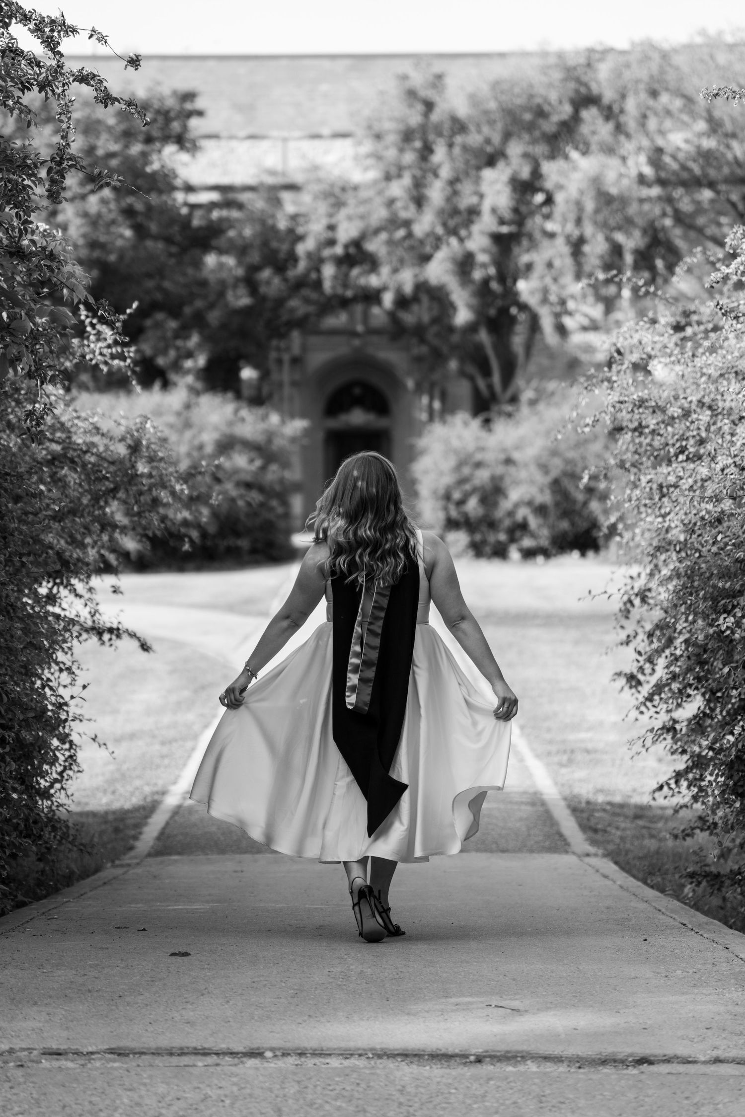 A black and white photo of graduate walking down path in flowing dress and graduation gown.