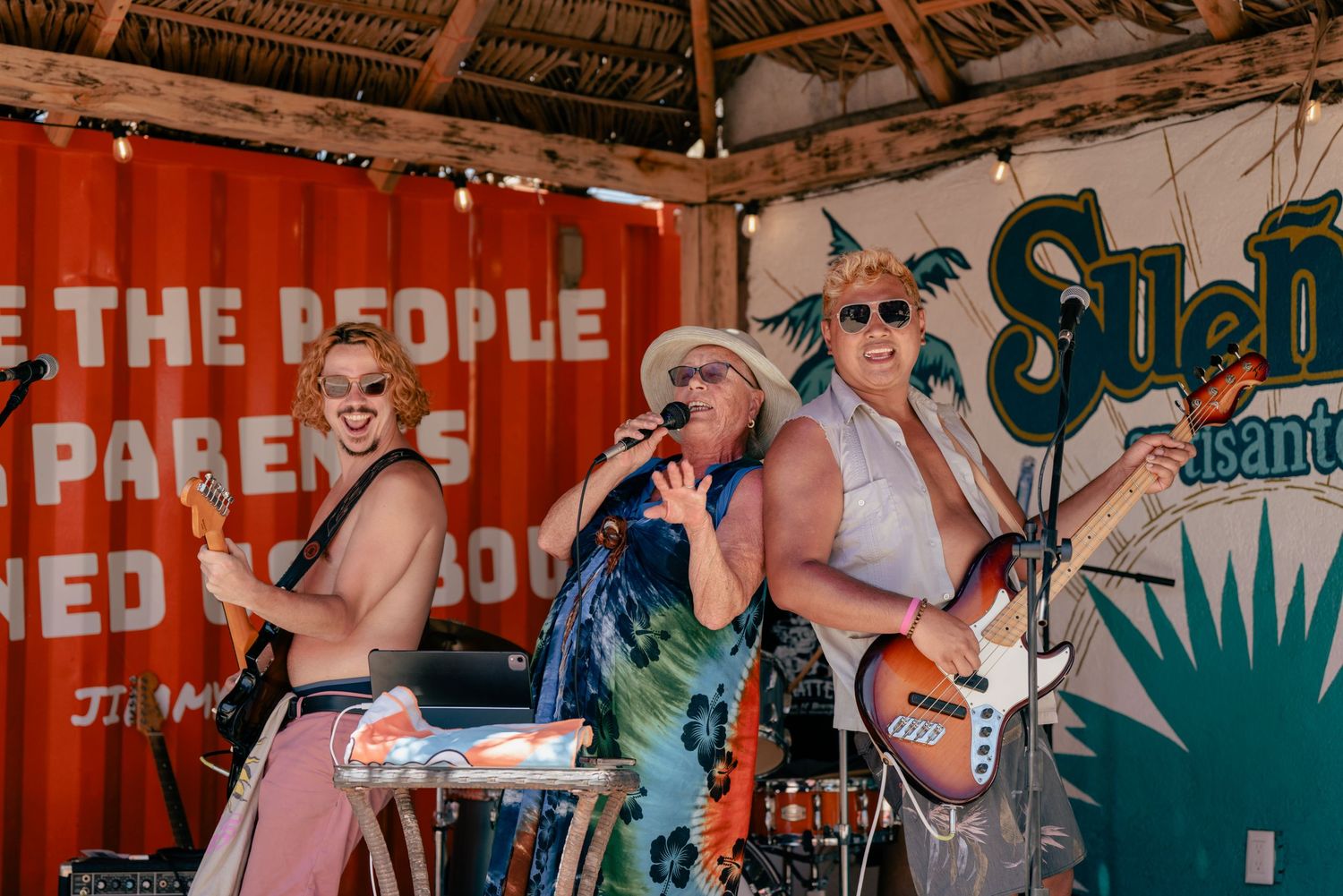 Musicians performing energetically on tropical-themed stage with red backdrop.