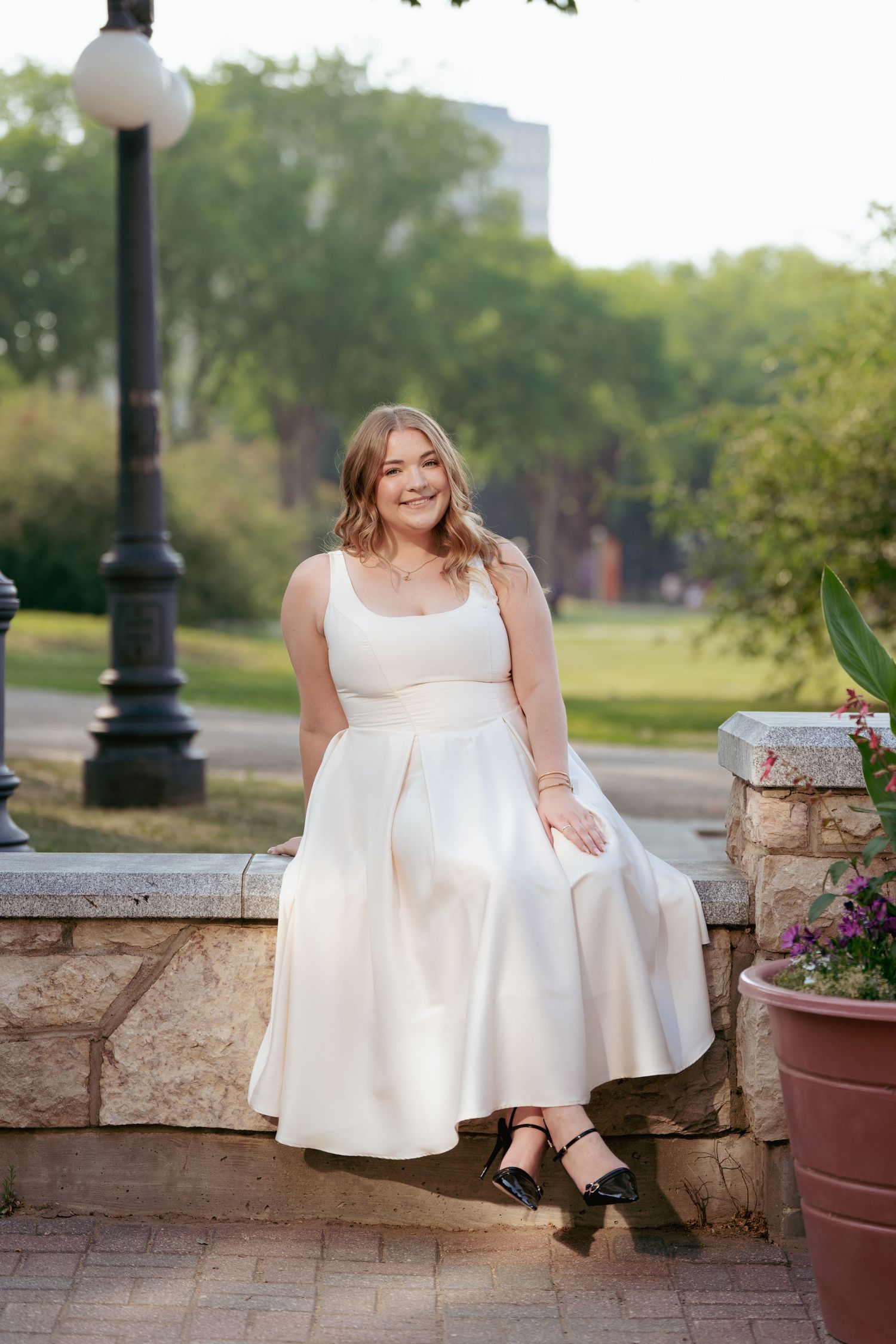 A graduate in elegant white dress poses on stone wall with lamp post and greenery.