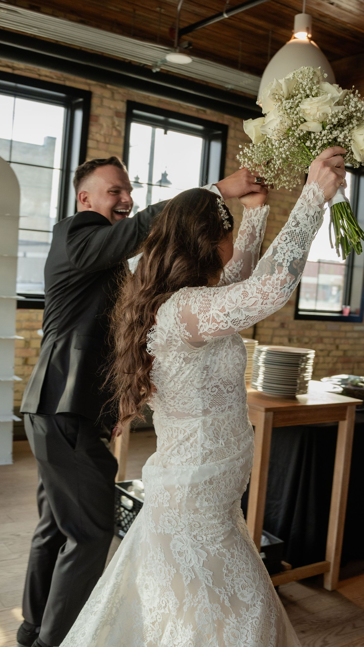 Wedding couple playfully reaching up together to grab hanging floral arrangement in industrial venue.