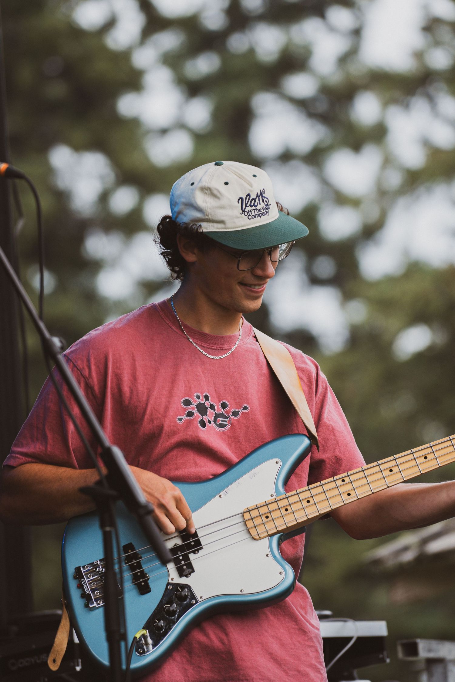 Musician playing light blue Fender bass guitar outdoors wearing a red t-shirt.