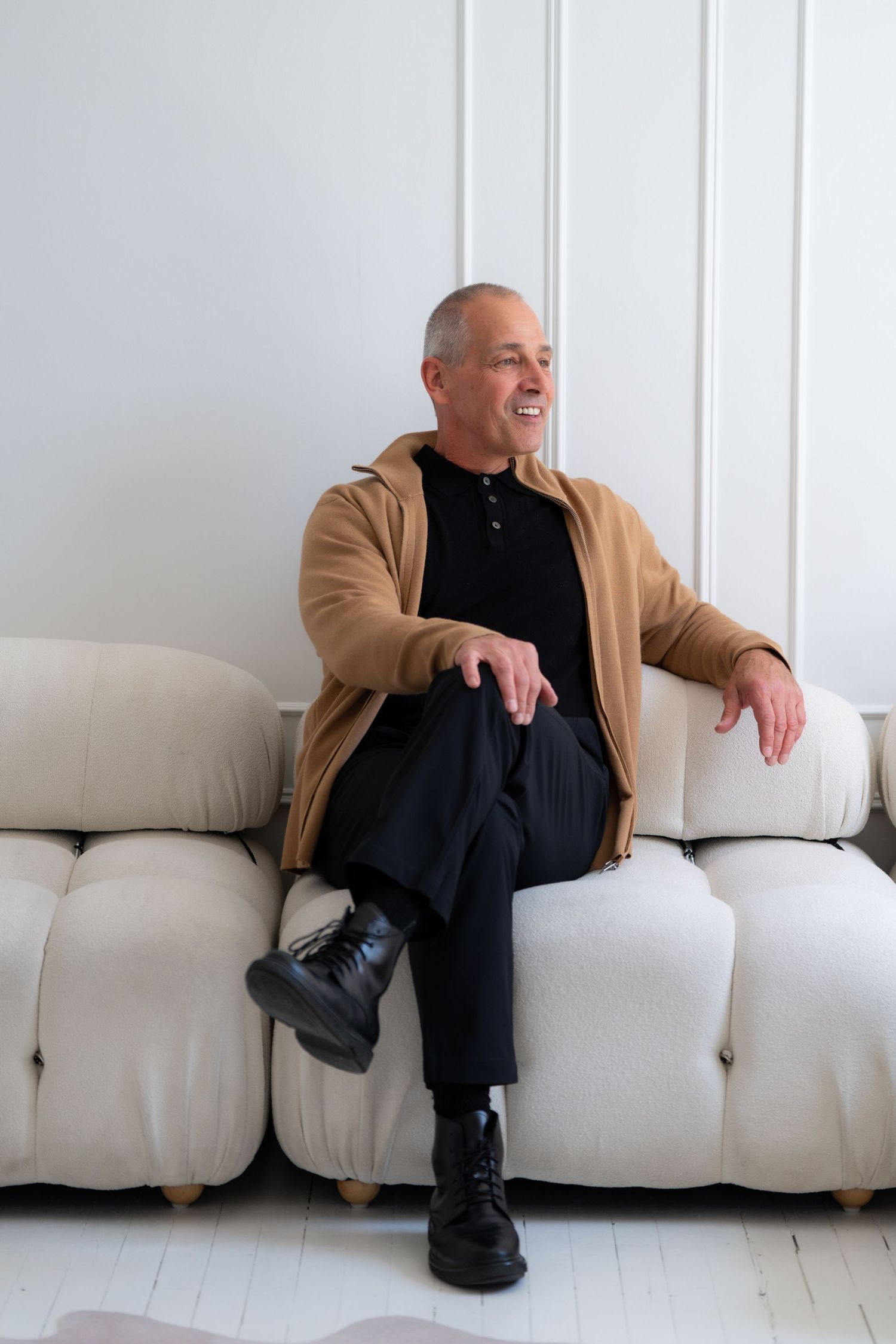 Professional studio photoshoot showing a person in black clothing and tan jacket sitting on a white couch.