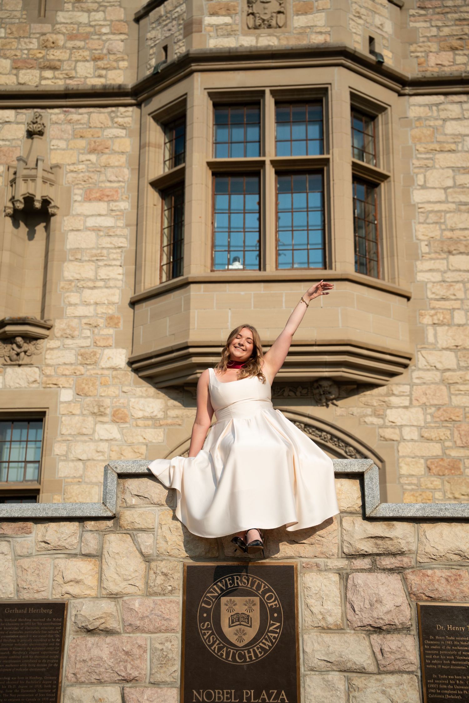A graduate in a white dress poses joyfully on a stone building ledge near an ornate bay window.
