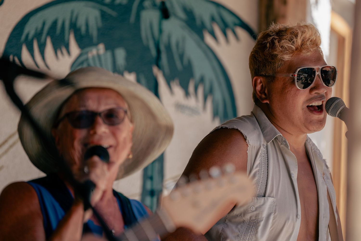 Musicians singing into microphones against palm tree mural backdrop in sunlit venue.