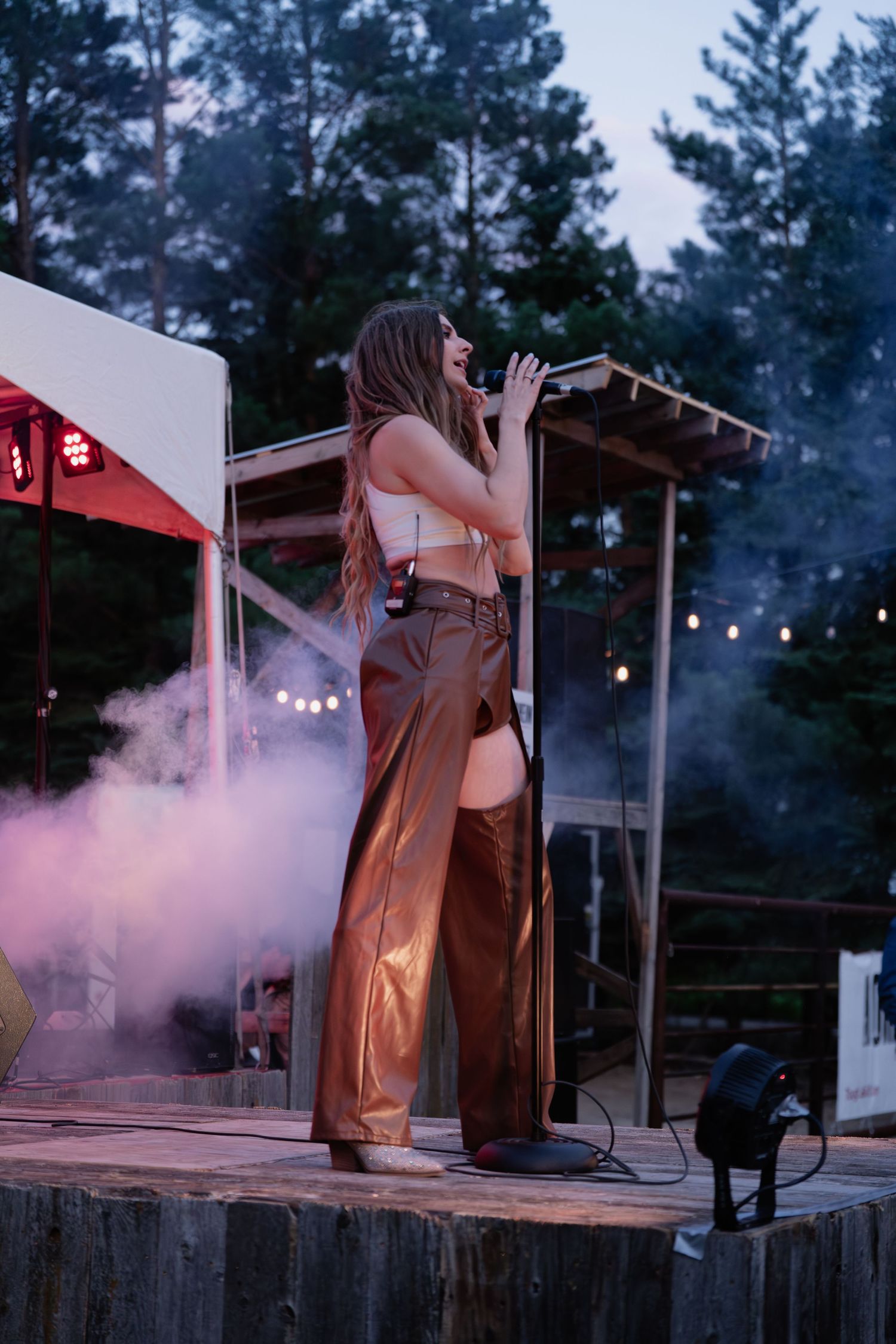 People performing on an outdoor stage with purple smoke effects and string lights at dusk.