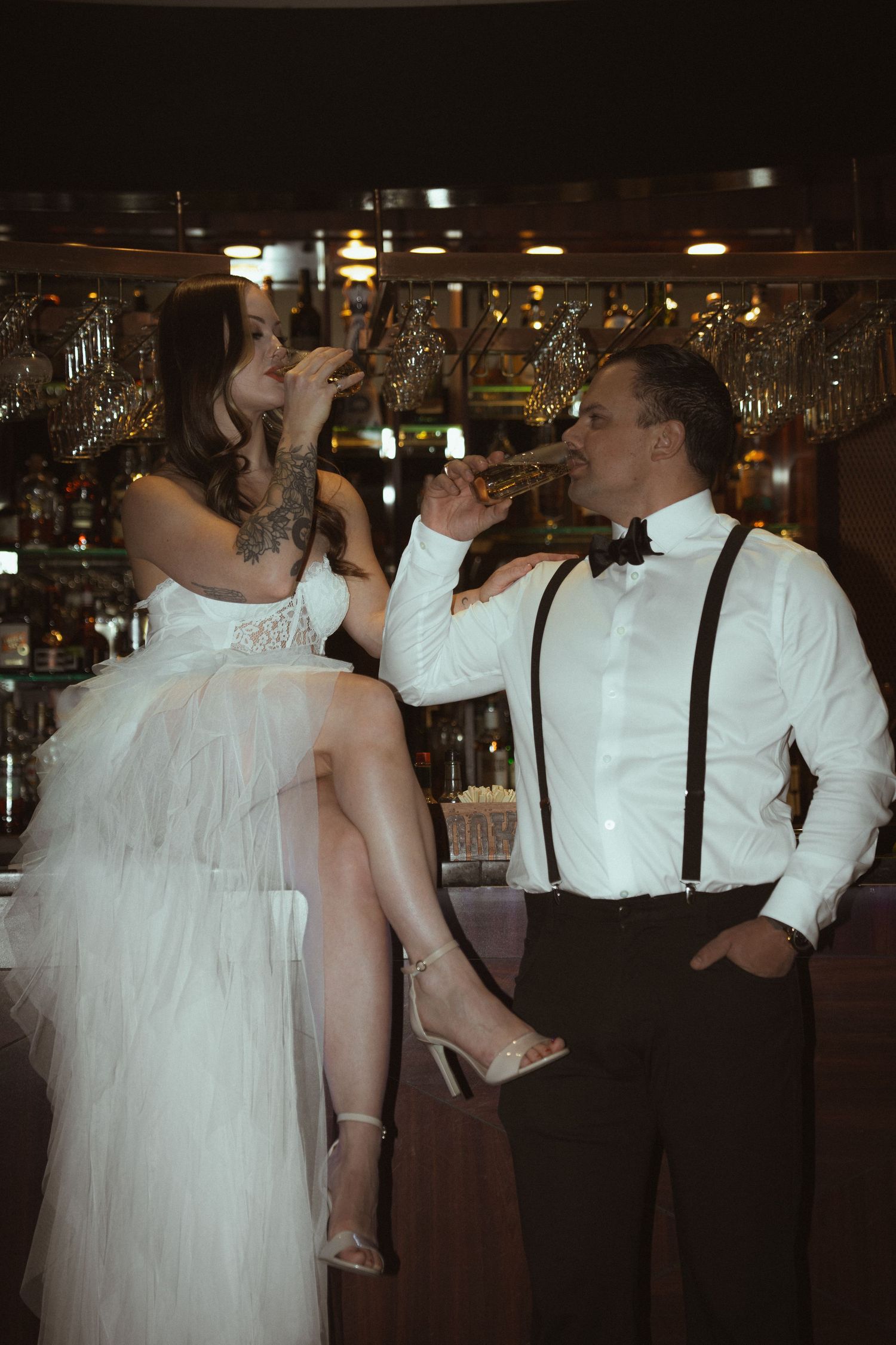 Wedding party members in white shirts and suspenders share drinks at a bar during celebration.