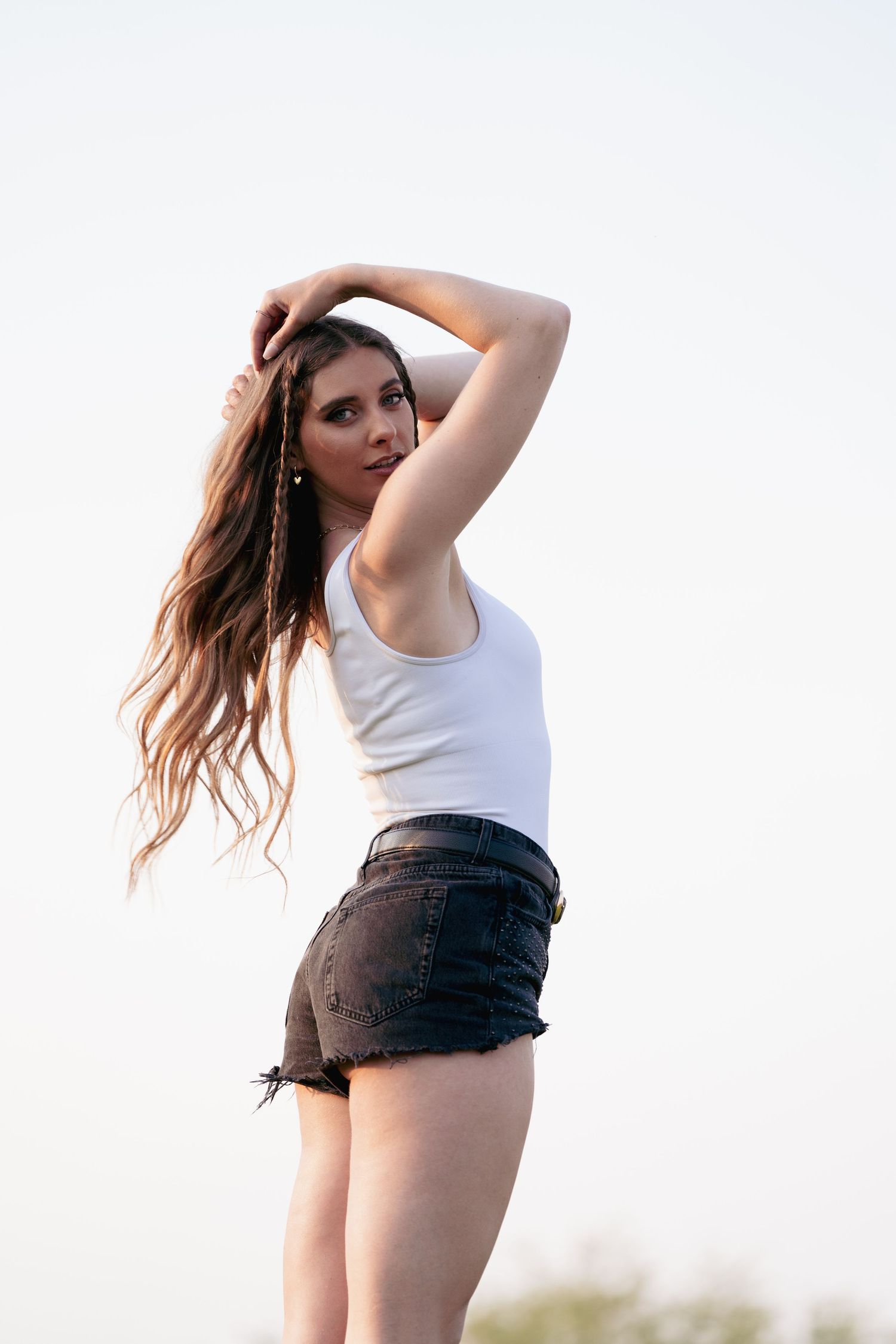 Young woman in denim shorts and white tank top poses against bright sky with flowing long hair.