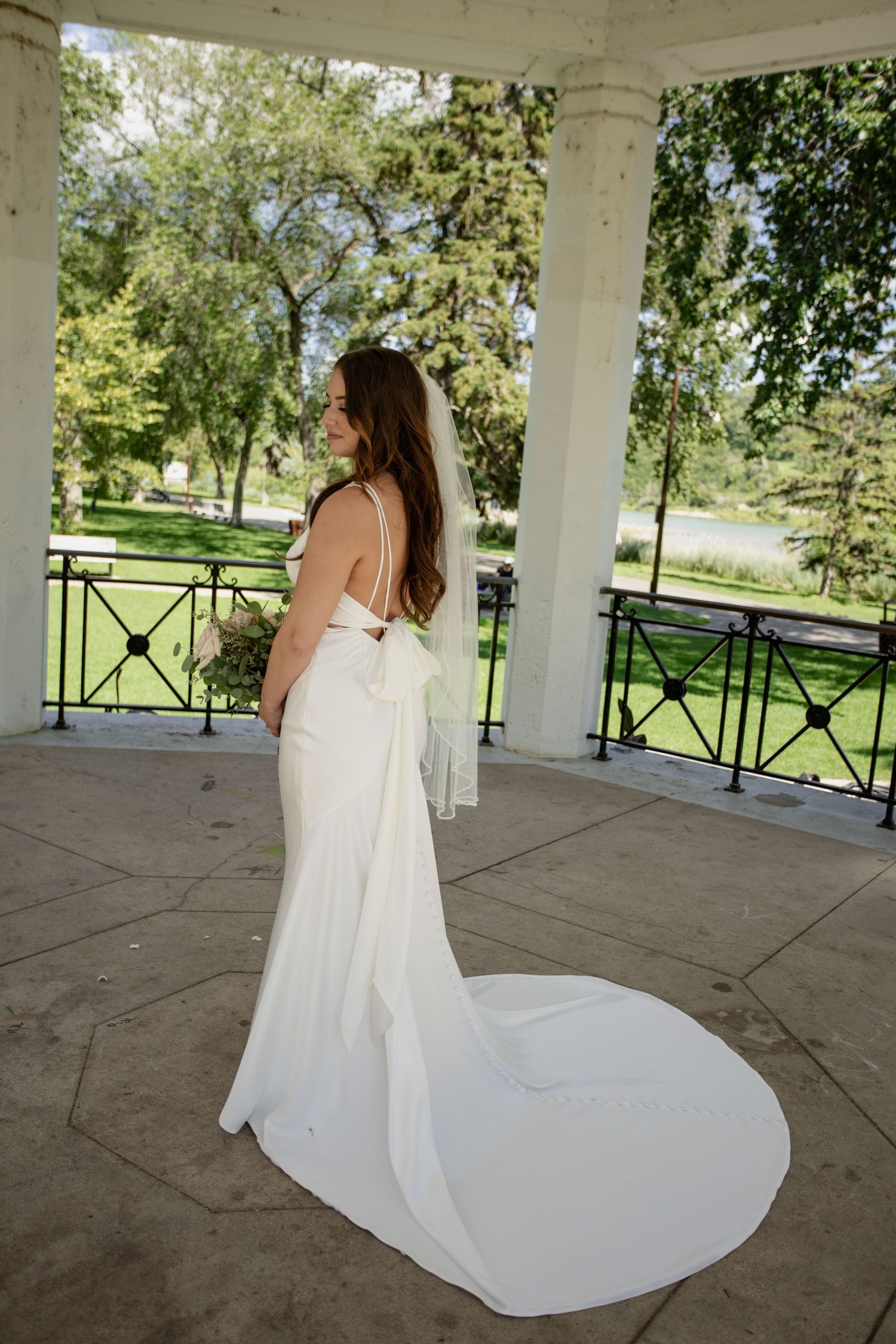 Bride in flowing white dress with train poses in outdoor gazebo.