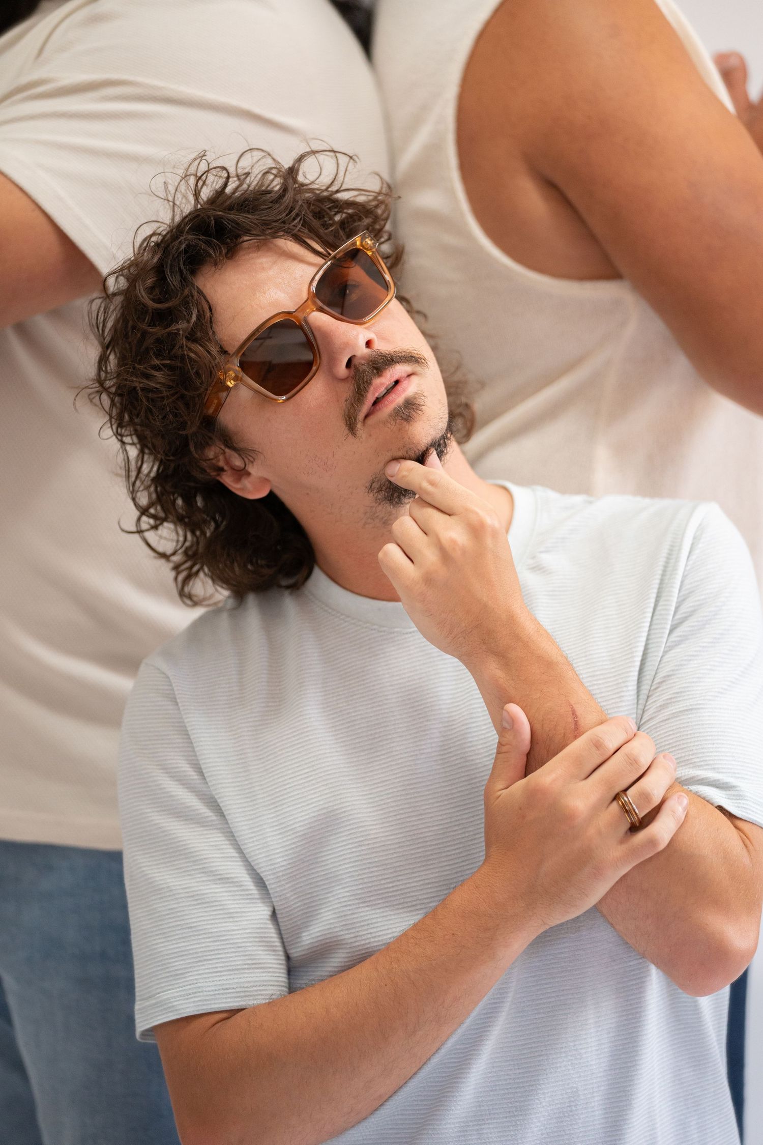 Casual portrait of a person in white t-shirt with curly hair wearing sunglasses and touching their chin.