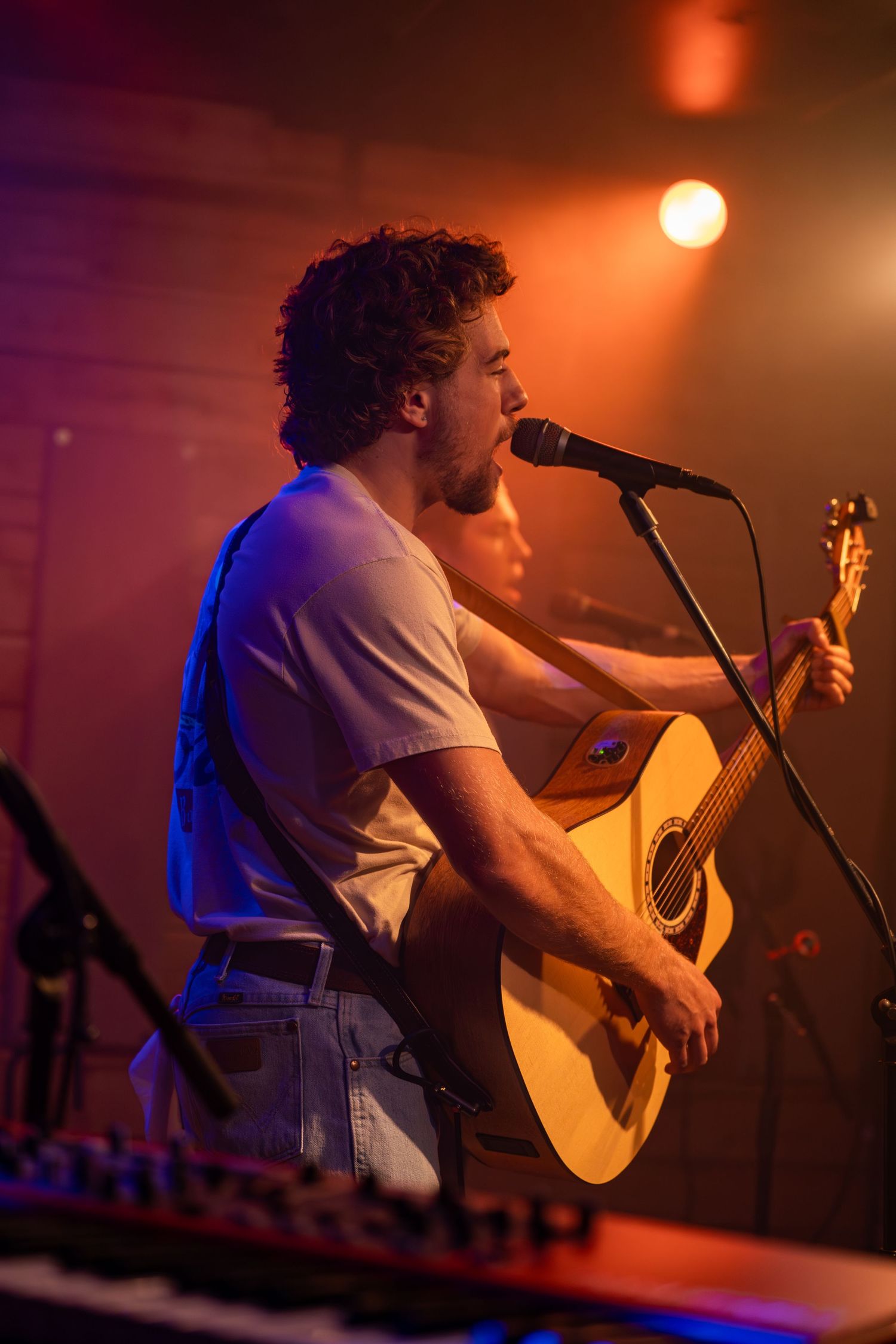 Musician performs on acoustic guitar under dramatic stage lighting with orange glow.