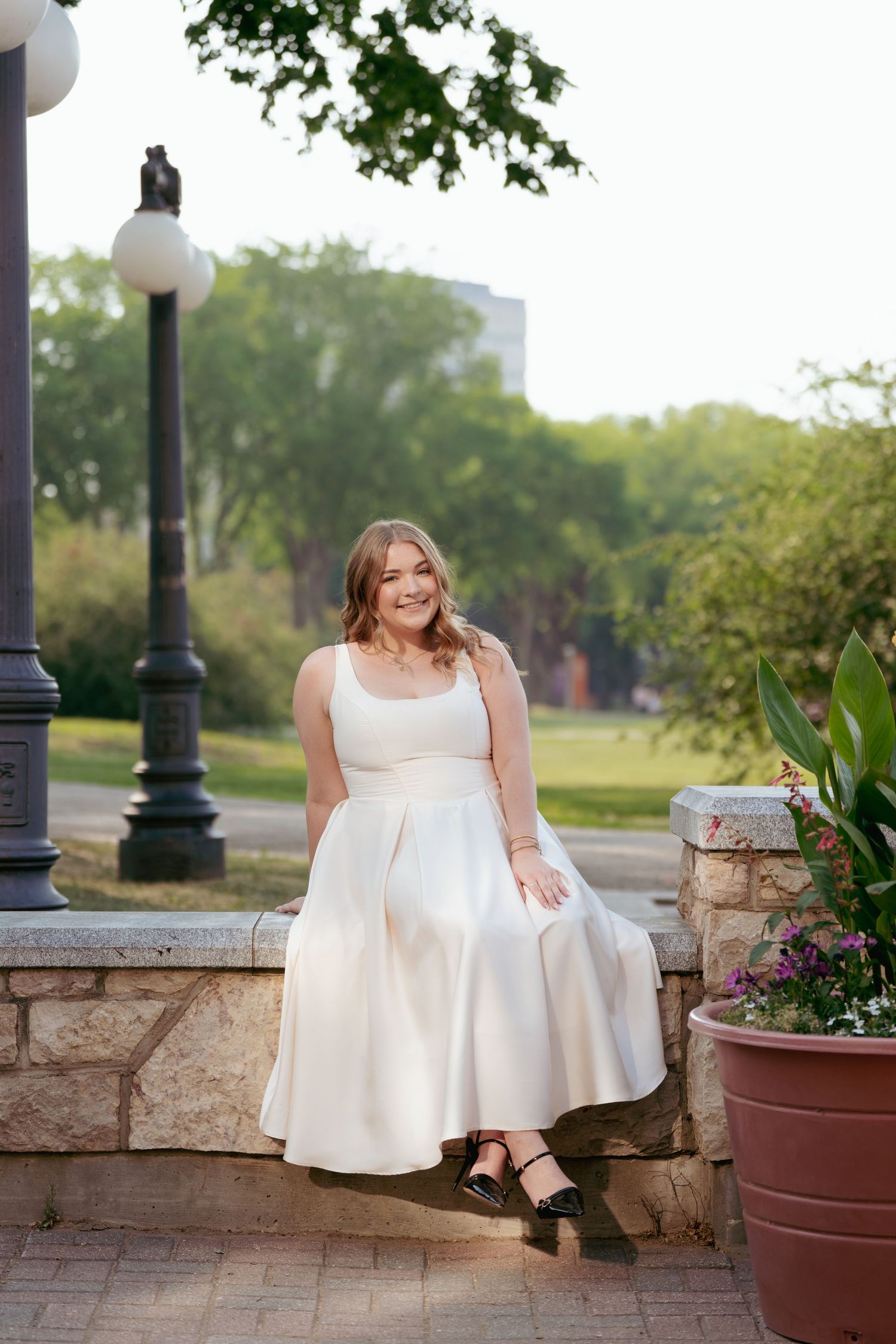 A graduate in white dress sits on stone wall near lamp post and flower pot.