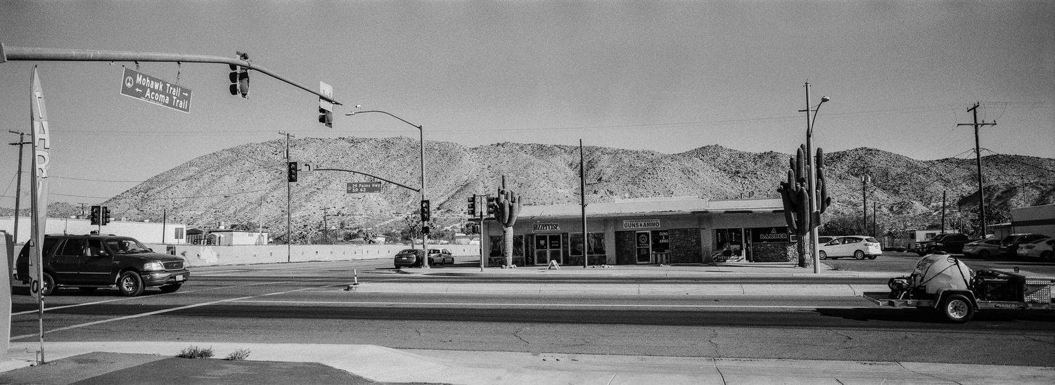 Black and white panoramic street scene with mountain backdrop, storefronts, traffic lights, and parked cars.